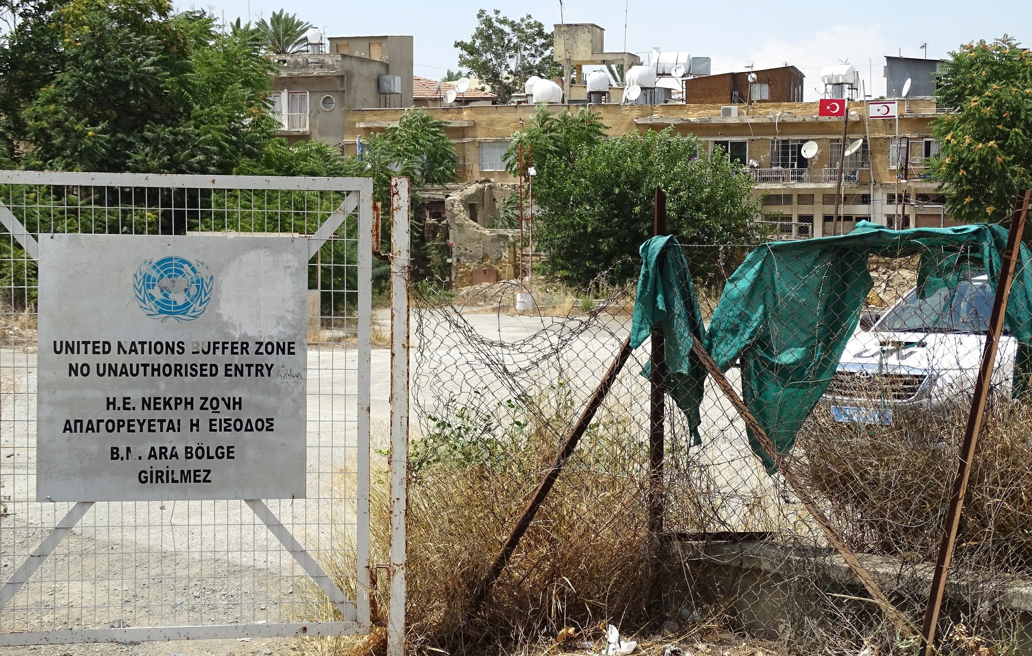 L'image montre une zone entourée d'une clôture. À gauche, il y a un panneau de la zone tampon des Nations Unies, indiquant qu'il s'agit d'un espace où l'entrée est interdite sans autorisation. À l'arrière-plan, on peut voir des bâtiments avec des antennes sur le toit, ainsi que de la végétation désordonnée au sol, créant une atmosphère de négligence. Le tout semble évoquer une région où l'accès est strictement contrôlé, probablement en raison de tensions historiques.