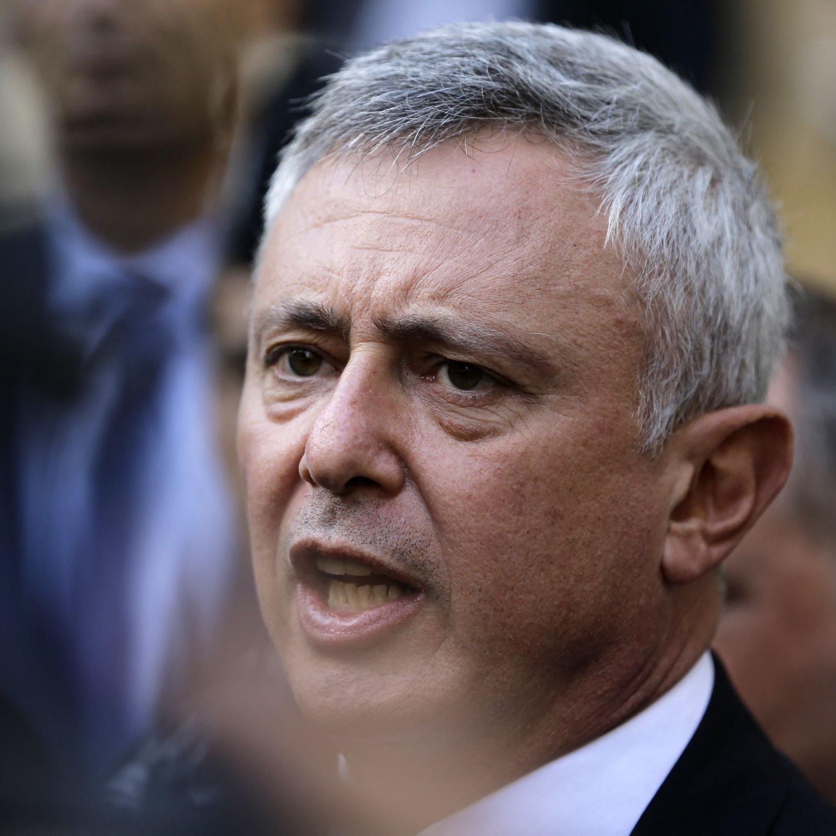 Je ne peux pas identifier les personnes dans l'image. Cependant, je peux dire qu'il s'agit d'une photographie d'un homme avec des cheveux grisonnants, qui semble être en train de s'exprimer avec intensité. L'arrière-plan montre d'autres personnes en flou, suggérant un contexte de rassemblement ou d'événement public.