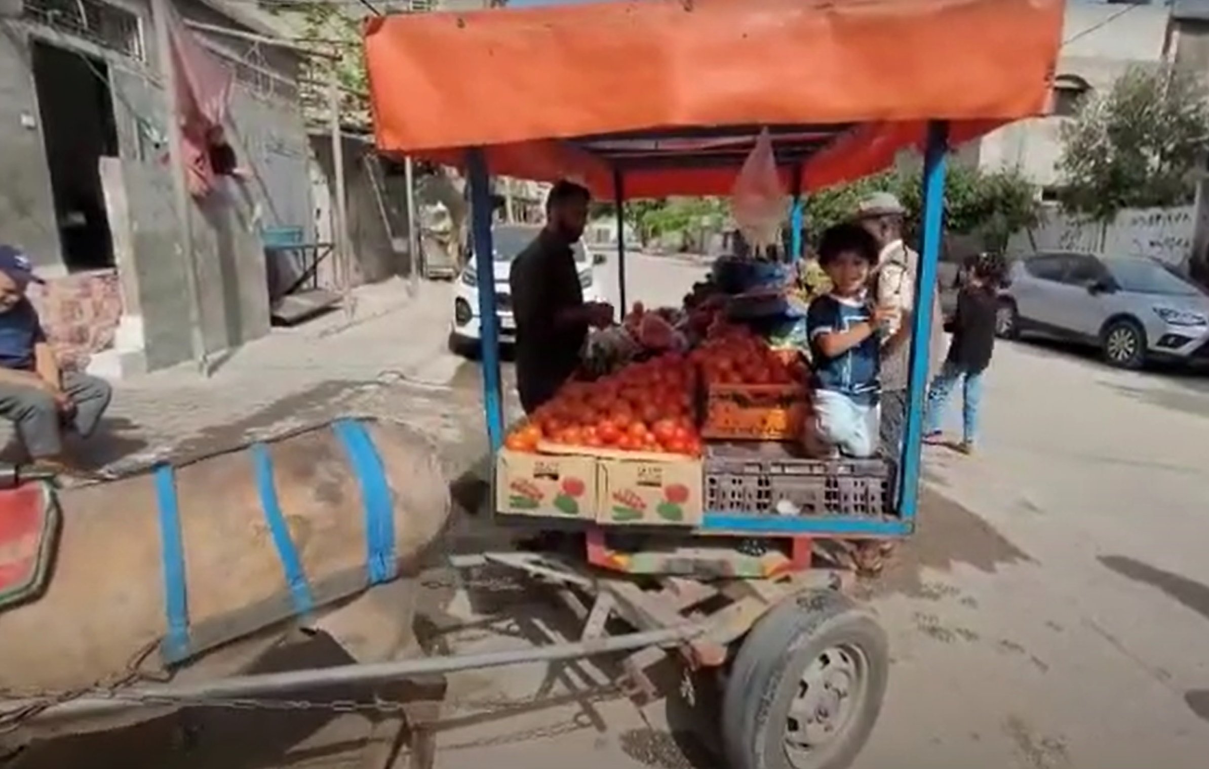 در این تصویر، یک دسته از میوه‌ها، به ویژه گوجه‌فرنگی، در یک وانت کوچک یا نیسان قرار دارد که در کنار خیابان پارک شده است. یک مرد در حال فروش میوه‌ها و یک کودک در کنار او به نظر می‌رسد. در گوشه تصویر، یک مرد دیگر در حال نشستن بر روی زمین است. خیابان و ساختمان‌های اطراف نیز در پس‌زمینه قابل مشاهده هستند. 