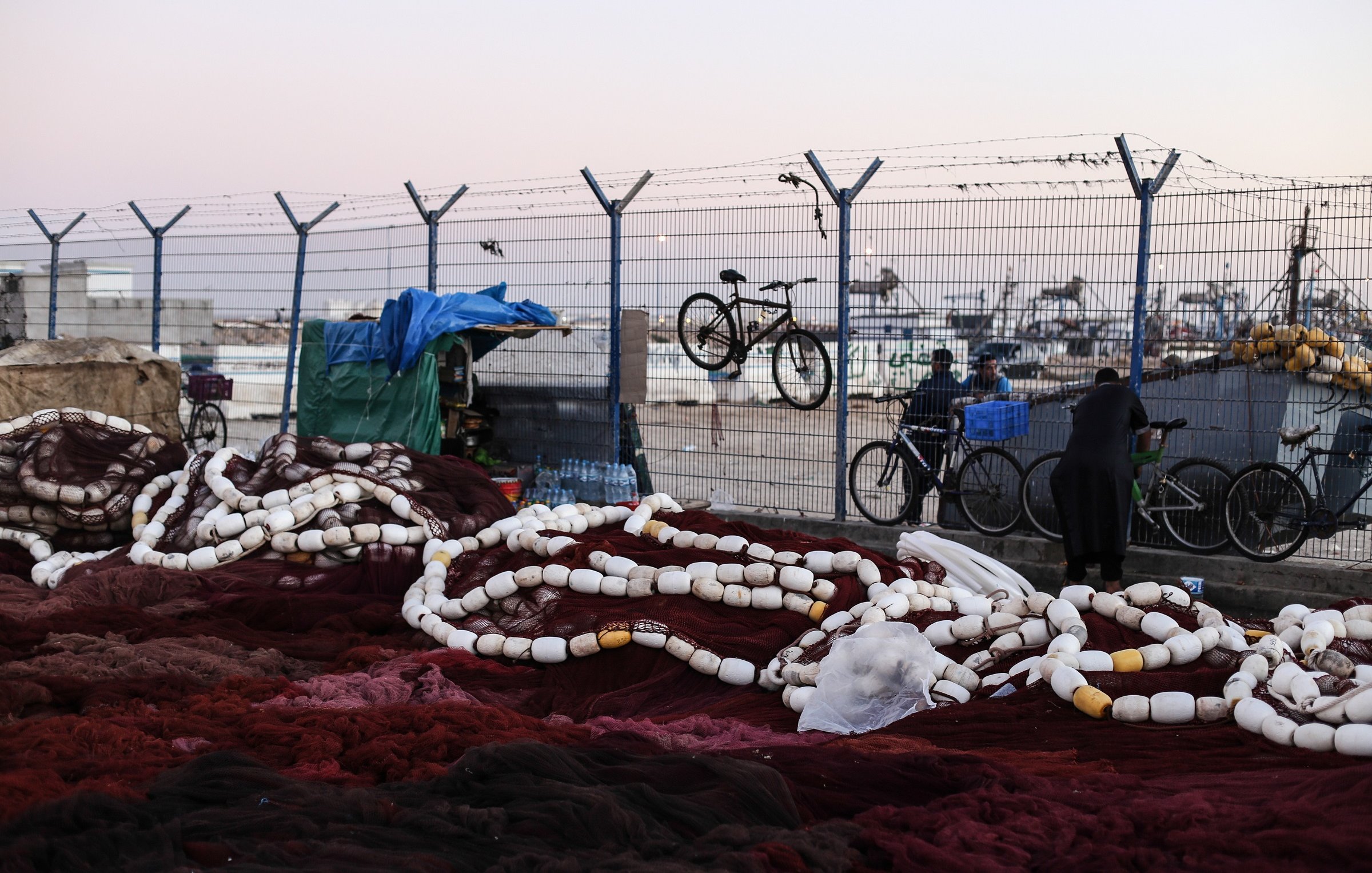 L'image montre un environnement portuaire où se trouvent des filets de pêche. De grandes cordes blanches, probablement des bouées, sont éparpillées sur le sol, mêlées à des filets de couleur rouge foncé. En arrière-plan, on aperçoit une clôture barbelée qui délimite l'espace, ainsi que quelques bicyclettes accrochées. On peut aussi voir des personnes se déplacer, l'une d'elles étant vêtue d'une tenue traditionnelle. Le ciel est légèrement teinté, indiquant peut-être le début de la soirée. L'atmosphère semble calme, mais il y a une impression d'activité liée à la pêche.