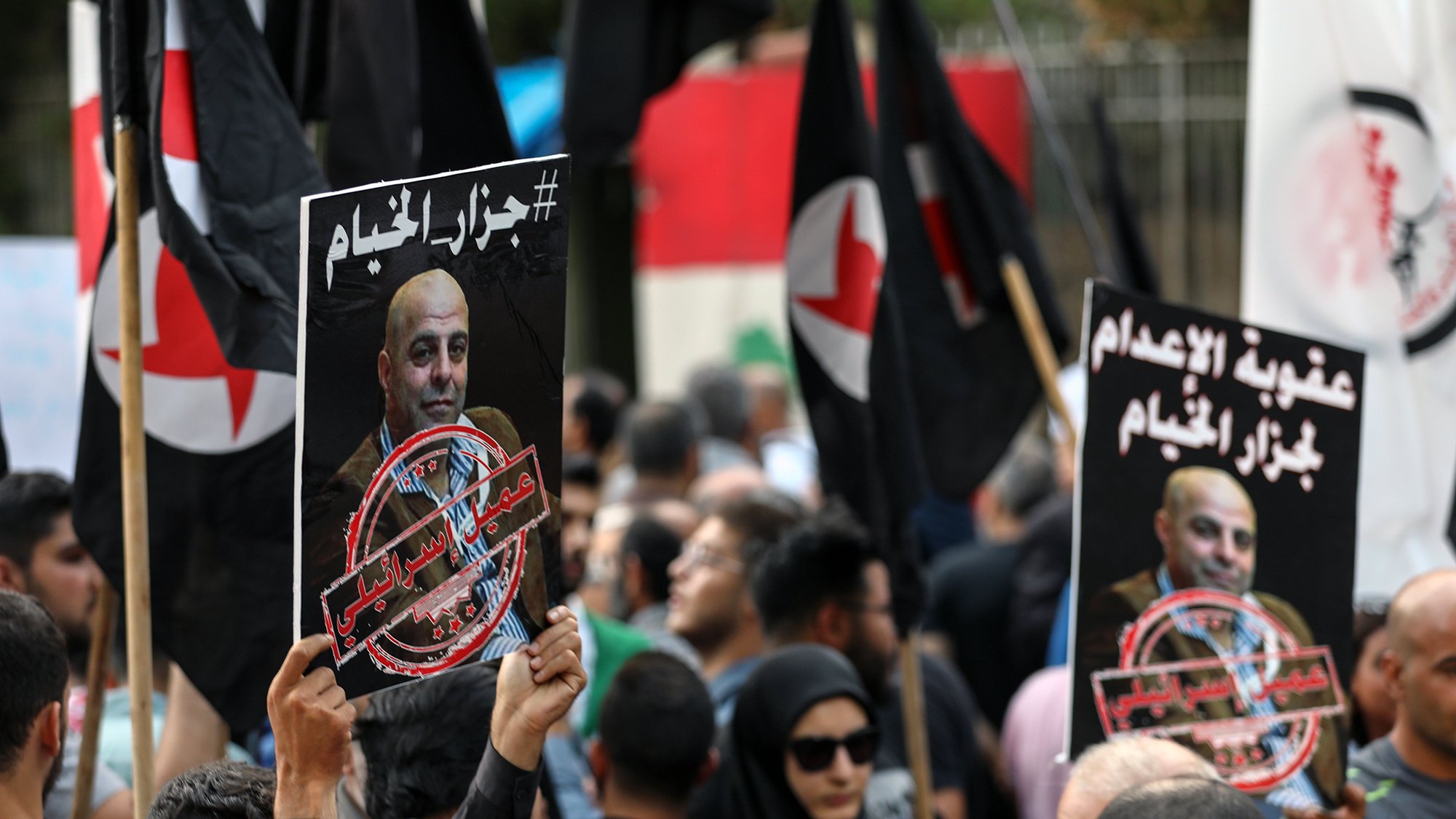 The image depicts a crowd of protesters holding signs and flags. The signs feature a portrait of a man along with a hashtag and phrases in Arabic, indicating a political or social issue. The crowd appears engaged and is likely part of a demonstration. The flags are predominantly black with red symbols, contributing to the atmosphere of the protest. The environment suggests a public gathering, possibly in response to a recent event or issue.