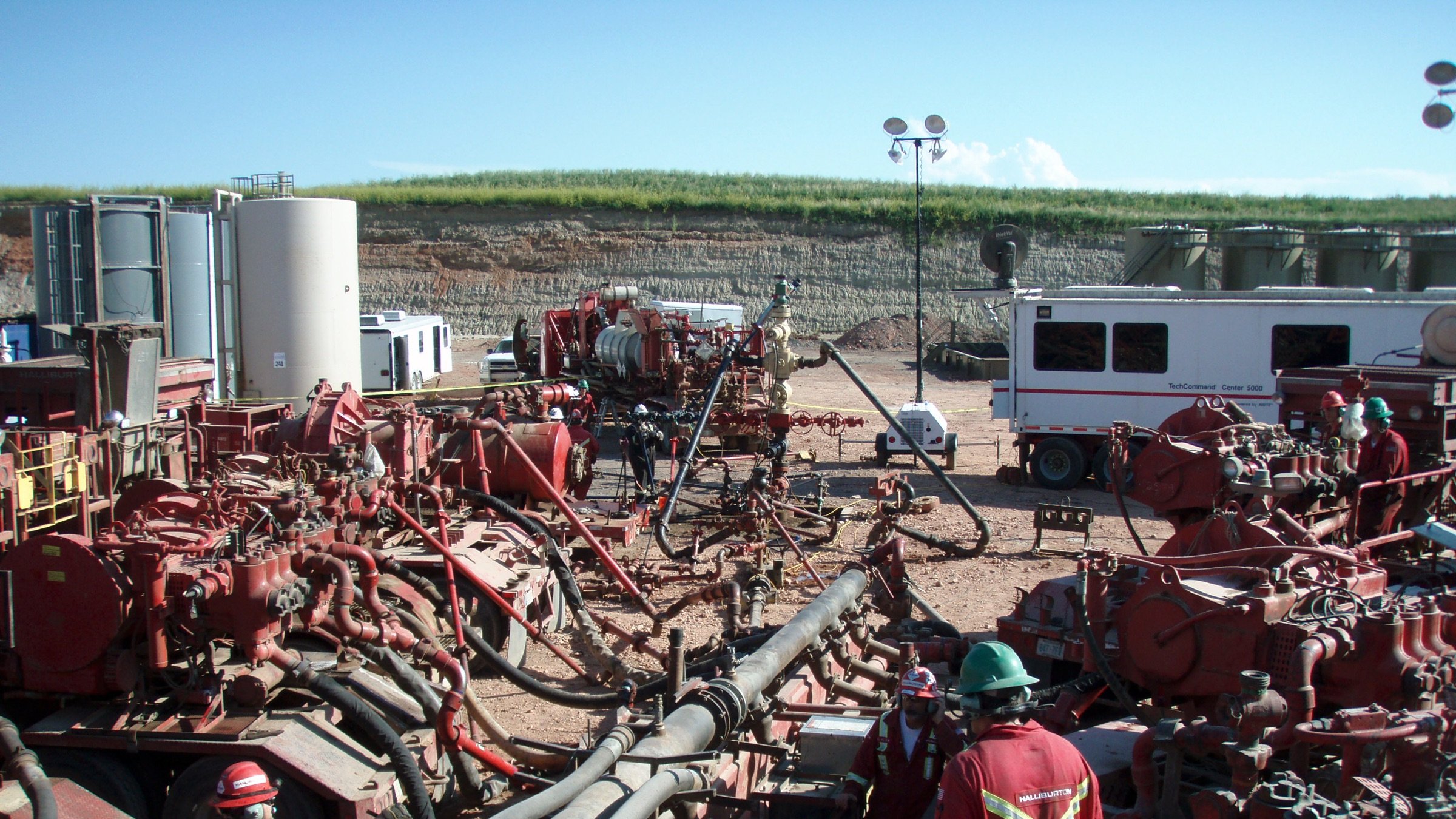 L'image montre un site d'exploitation, probablement lié à l'industrie pétrolière ou gazière. On y voit plusieurs équipements imposants, notamment des pompes et des tuyaux, ainsi que des travailleurs en tenue de sécurité, portant des casques et des vêtements rouges. Le paysage en arrière-plan est rural, avec une colline et des installations industrielles. Le ciel est dégagé, laissant suggérer une journée ensoleillée. L'ensemble de la scène évoque une activité intense et technique, généralement associée à des opérations d'extraction ou de forage.