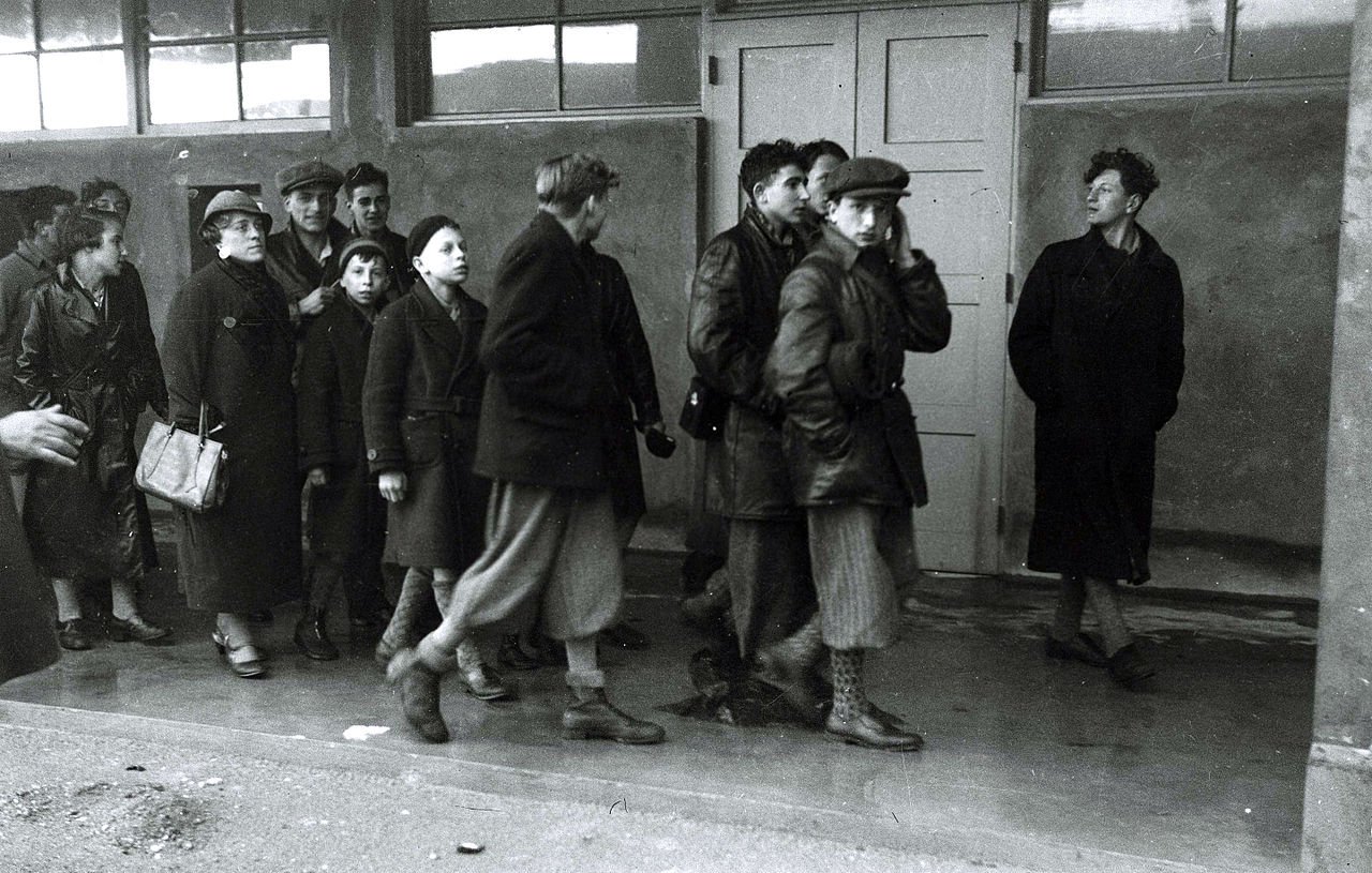 این تصویر سیاهوسفید گروهی از افراد را نشان میدهد که در یک محیط داخلی در حال حرکت هستند. برخی از افراد لباسهای گرم و کلوفت به تن دارند و به نظر میرسد که در حال عبور از یک فضای بسته هستند. تعدادی از افراد در حال گفتگو یا مراقب هستند، در حالی که دیگران به جلو نگاه میکنند. فضای تصویر حالت جدی و متفکری دارد و به طور کلی جو نگرانکنندهای را منتقل میکند.
