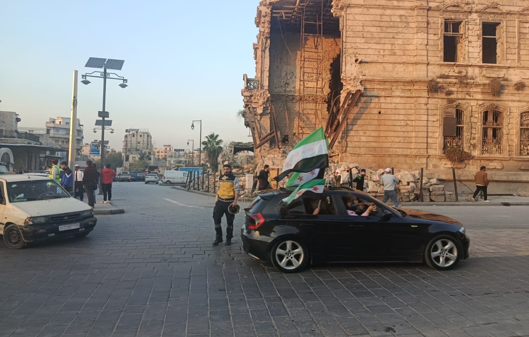Scène de rue avec un bâtiment endommagé, des gens et une voiture avec un drapeau.