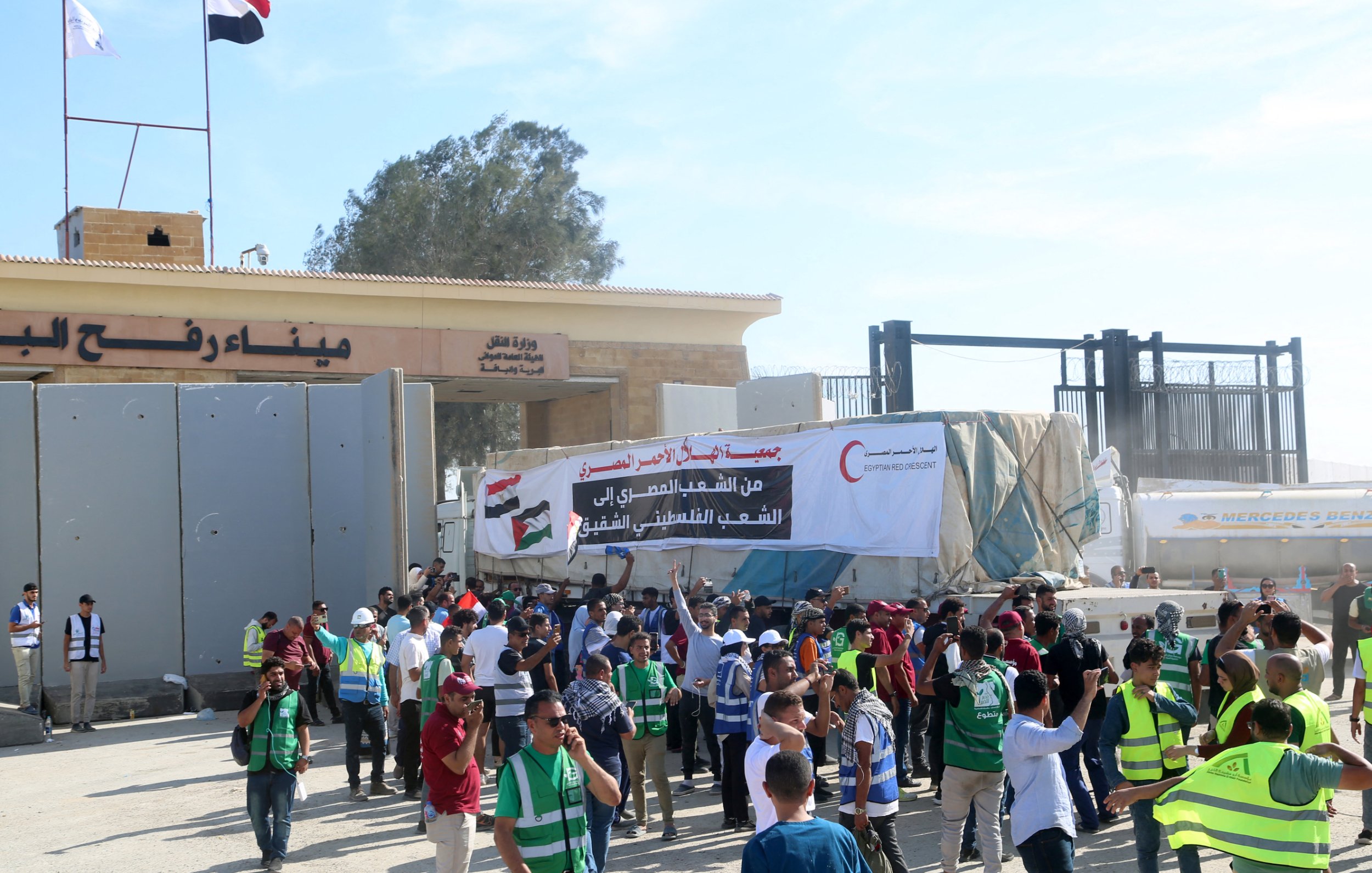 L'immagine mostra un gruppo di persone radunate davanti a un'entrata o a un confine, con bandiere nazionali visibili. Ci sono manifestanti che indossano gilet verdi e alcuni stanno tenendo striscioni. L'atmosfera sembra essere di protesta o di celebrazione, con diversi individui che interagiscono tra loro e partecipano all'evento. Sullo sfondo si vedono strutture architettoniche e una grande scritta in arabo.