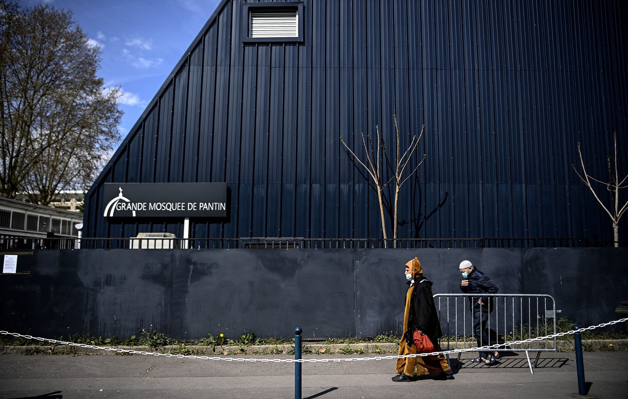 L'image montre une scène extérieure près d'un bâtiment. On peut voir la façade d'une grande mosquée, identifiée par un panneau indiquant "Grande Mosquée de Pantin". À l'avant-plan, deux personnes marchent le long d'un chemin, l'une d'elles portant un voile et une tenue traditionnelle, tandis que l'autre est vêtue d'une manière plus décontractée, portant également un masque. L'architecture du bâtiment est moderne, avec un toit incliné, et l'environnement semble urbain, avec quelques arbres dépouillés de feuilles. Une barrière de sécurité sépare la rue du bâtiment.
