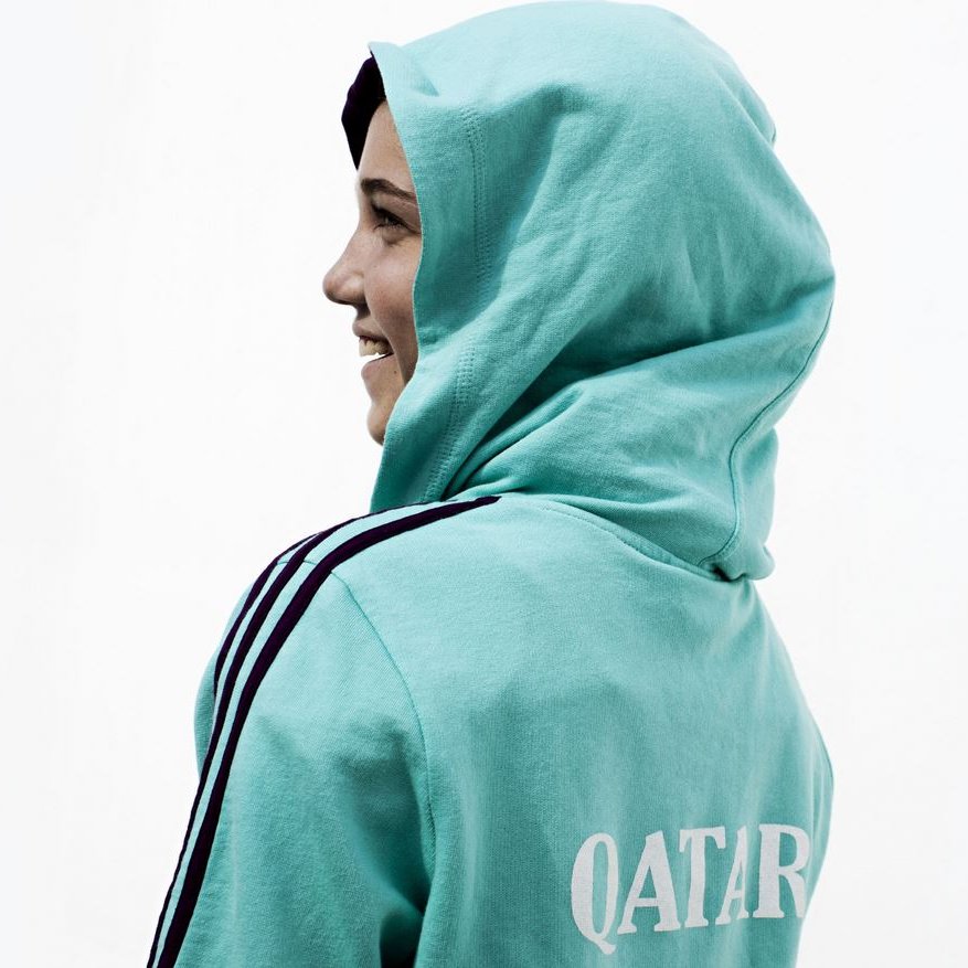 L'image montre une personne de profil, portant un sweat à capuche de couleur turquoise. Le sweat arbore le mot "QATAR" inscrit à l'arrière. La personne semble sourire, et la capuche est relevée sur sa tête. L'arrière-plan est neutre, mettant l'accent sur la tenue de la personne.