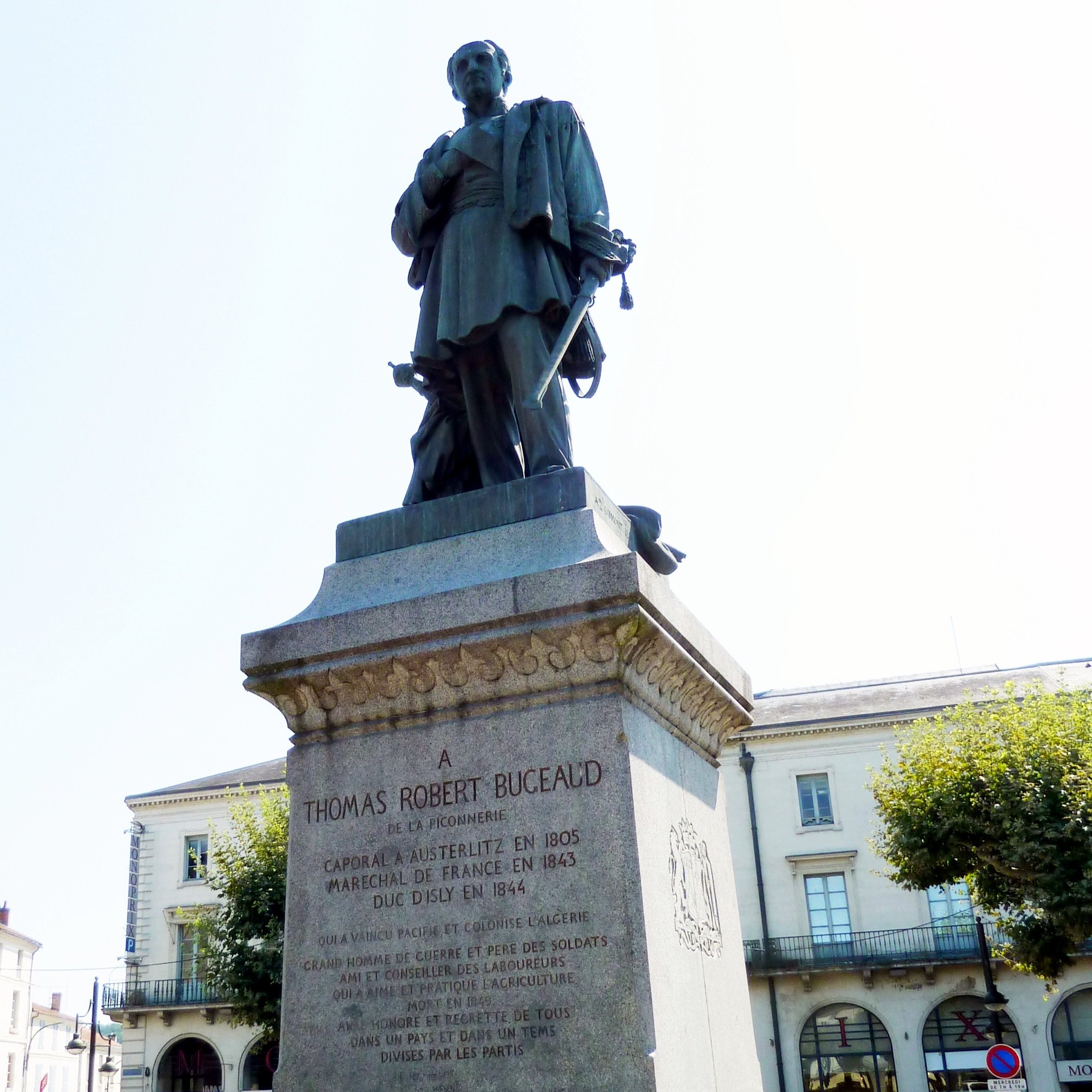 L'image représente une statue érigée à la mémoire de Thomas Robert Bugeaud. La statue est placée sur un socle en pierre portant une inscription détaillant ses accomplissements. Bugeaud est présenté en tenue militaire, tenant son épée. En arrière-plan, on peut voir une rue avec des arbres et des bâtiments, indiquant probablement un espace public. La lumière est claire, suggérant une journée ensoleillée.