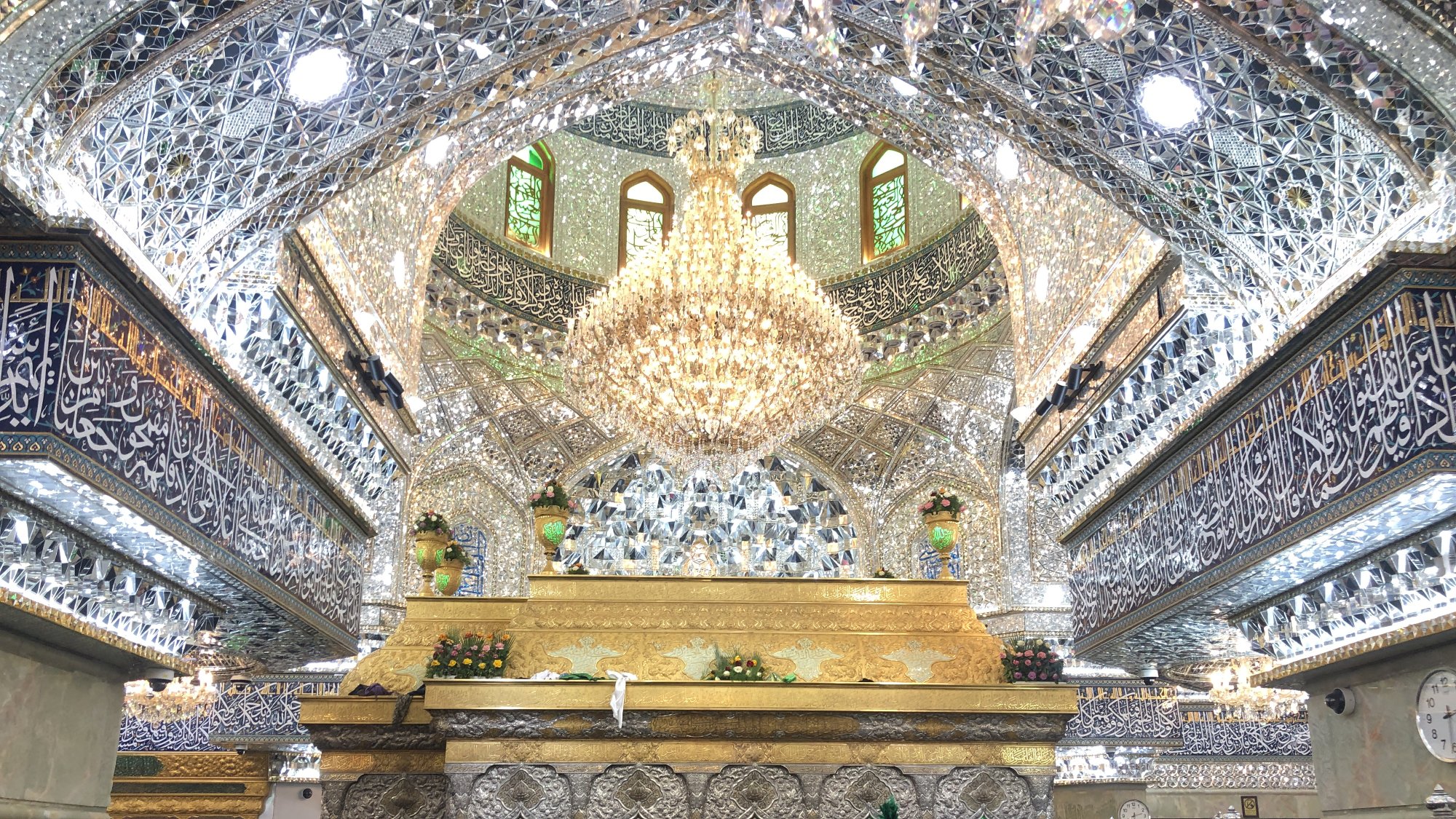 The image depicts the interior of a beautifully ornate religious site, characterized by intricate mirror work and elaborate decorations. The ceiling is adorned with shimmering tiles and a large chandelier, creating a luminous atmosphere. There is a golden structure at the center, likely a shrine or tomb, surrounded by a crowd of women wearing traditional black clothing, all appearing to be engaged in prayer or reflection. The walls and ceiling are richly decorated with calligraphy and other designs, enhancing the sacred ambiance of the space.