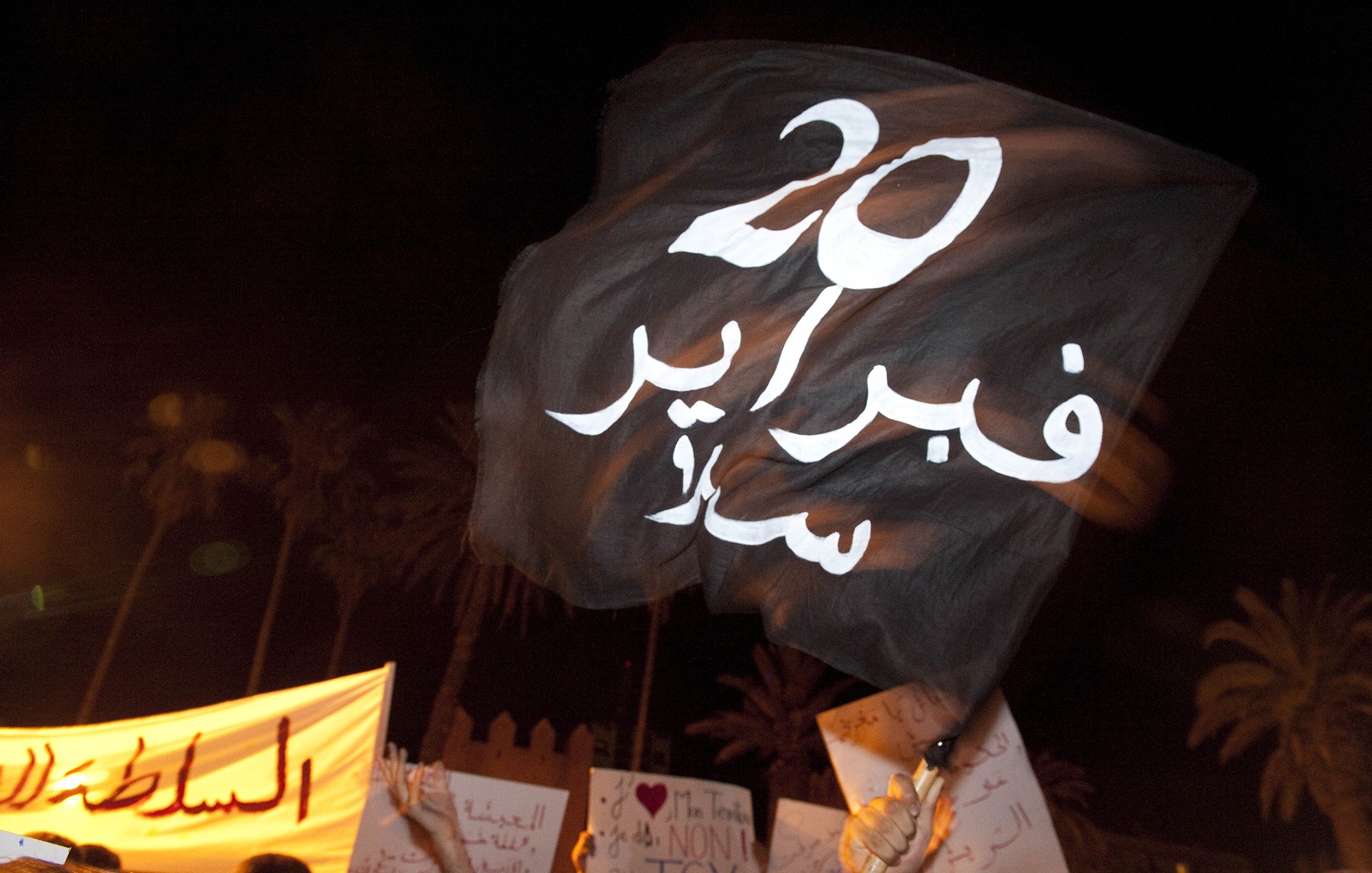 الصورة توضح لافتة تحمل علمًا أسود مكتوبًا عليه "20 فبراير" باللون الأبيض، وهو يوم معروف للاحتجاجات في المغرب. في الخلفية، يمكن رؤية لافتات أخرى تشير إلى احتجاجات جماهيرية. الأجواء تبدو حماسية، حيث يعكس المشهد نشاطًا سياسيًا واجتماعيًا.