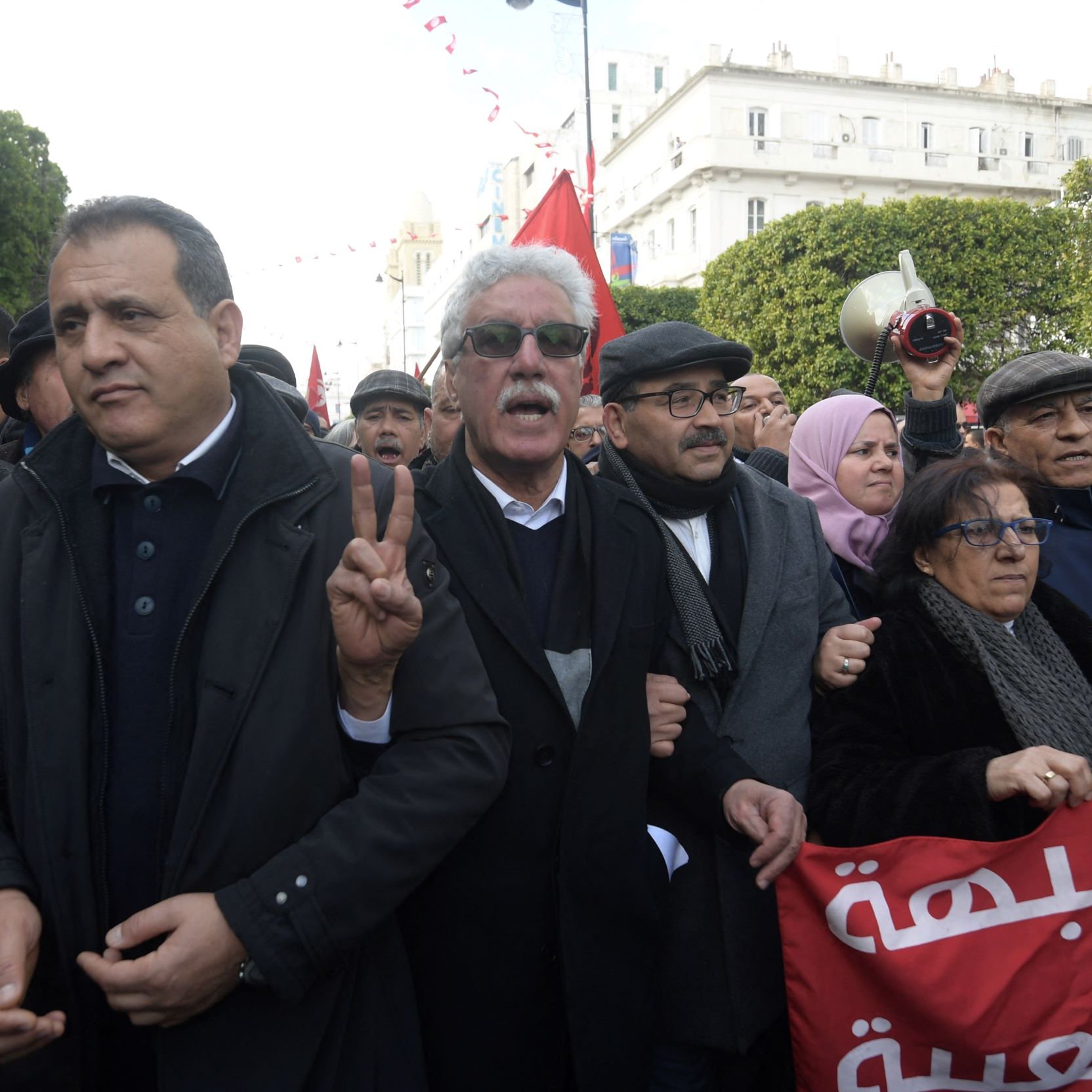 L'image montre un groupe de personnes rassemblées lors d'une manifestation. Au premier plan, on peut voir des hommes et des femmes exprimant des émotions vives, certains levant des signes de paix avec leurs mains. Les participants portent des vêtements variés, incluant des manteaux et des chapeaux. Des banderoles ou des drapeaux en arrière-plan ajoutent de la couleur à la scène, et l'atmosphère semble dynamique et engagée. Le décor environnant suggère un cadre urbain.