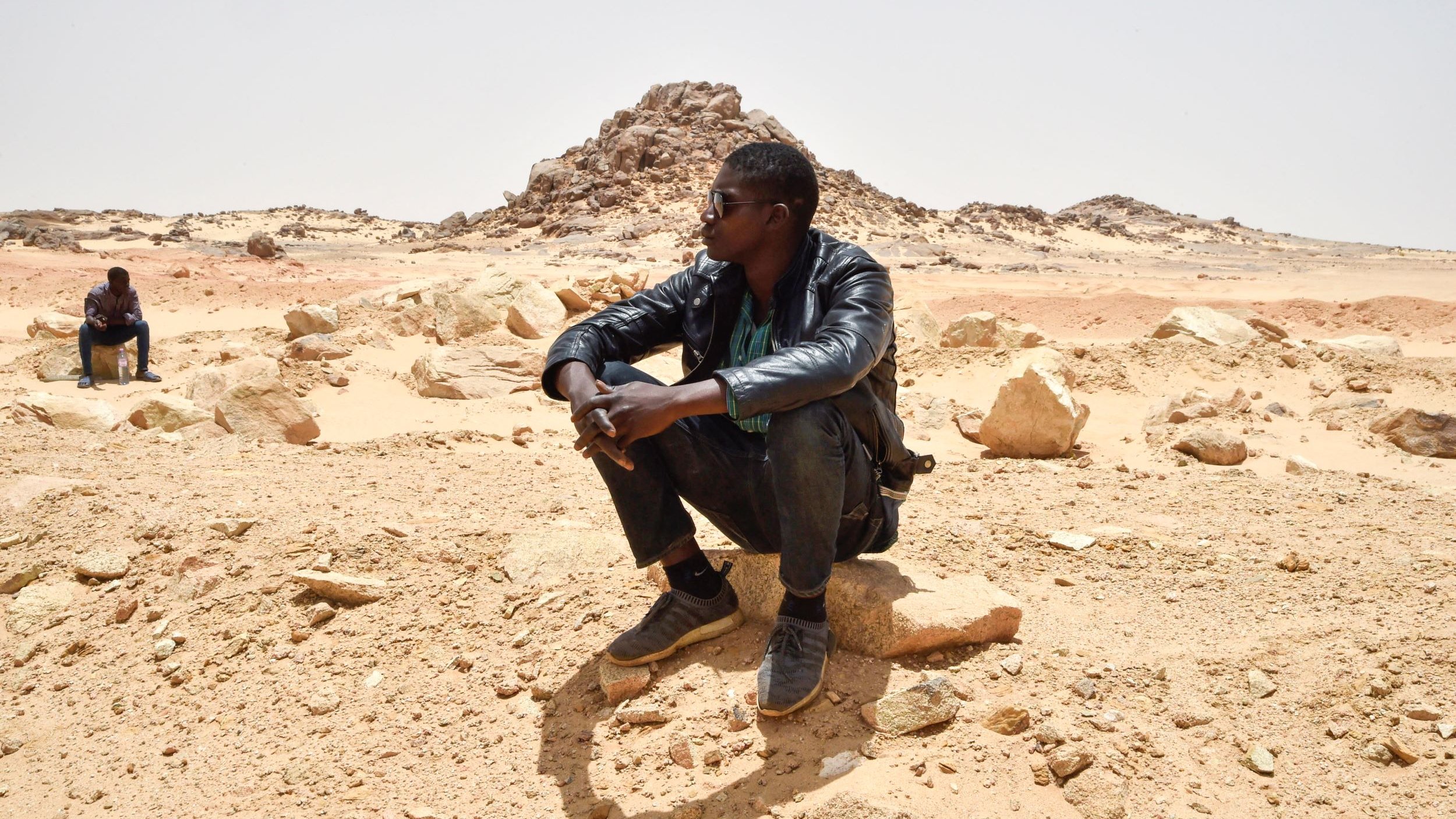L'image montre deux personnes assises dans un paysage désertique. L'un des hommes est en train de s'asseoir sur une grosse pierre, portant une veste en cuir et des lunettes de soleil. Il semble pensif, observant le paysage autour de lui. En arrière-plan, une autre personne est également assise, mais davantage éloignée. Le terrain est constitué de sable et de roches, avec des formations rocheuses au loin, sous un ciel clair. L'atmosphère semble calme et isolée.