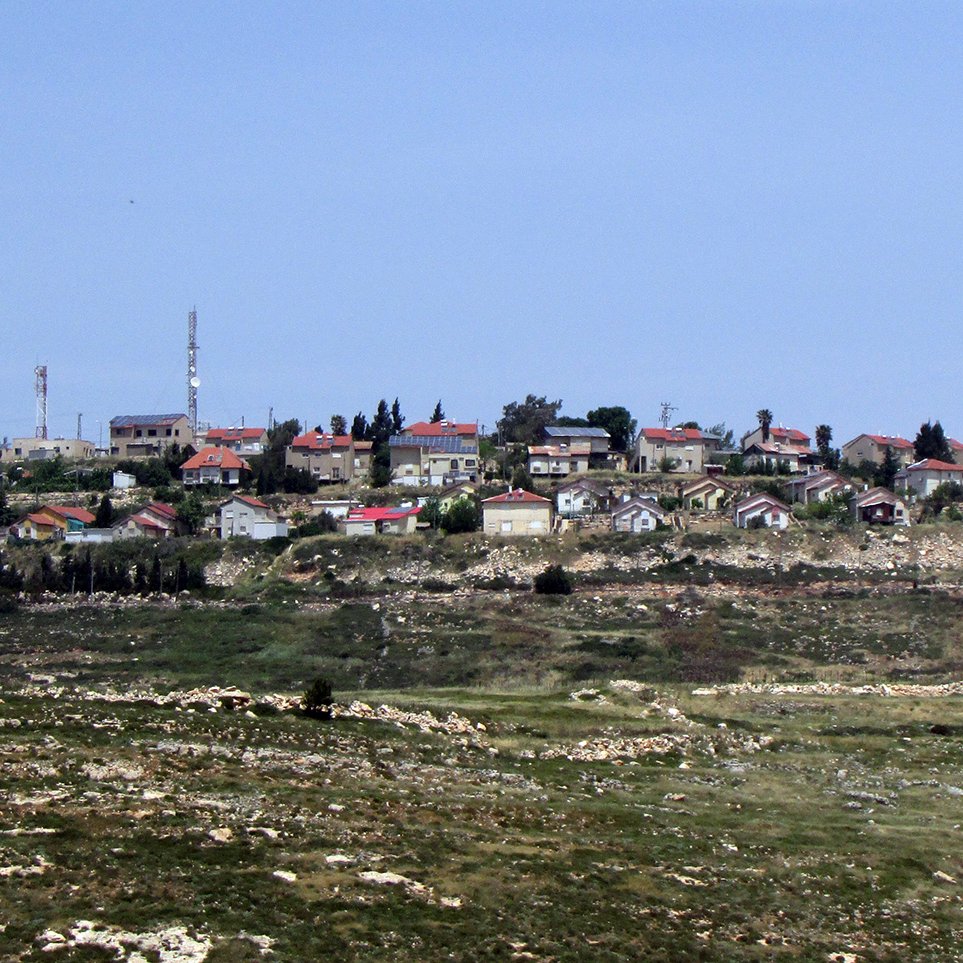 The image depicts a small village or settlement situated on a hillside. The buildings are primarily residential, featuring a mix of styles with terracotta roofs and natural stone walls. The landscape surrounding the village is hilly and green, with patches of rocky terrain and sparse vegetation. In the distance, there are communication towers and possibly other structures, indicating some level of infrastructure. The sky above is clear, suggesting a bright day.