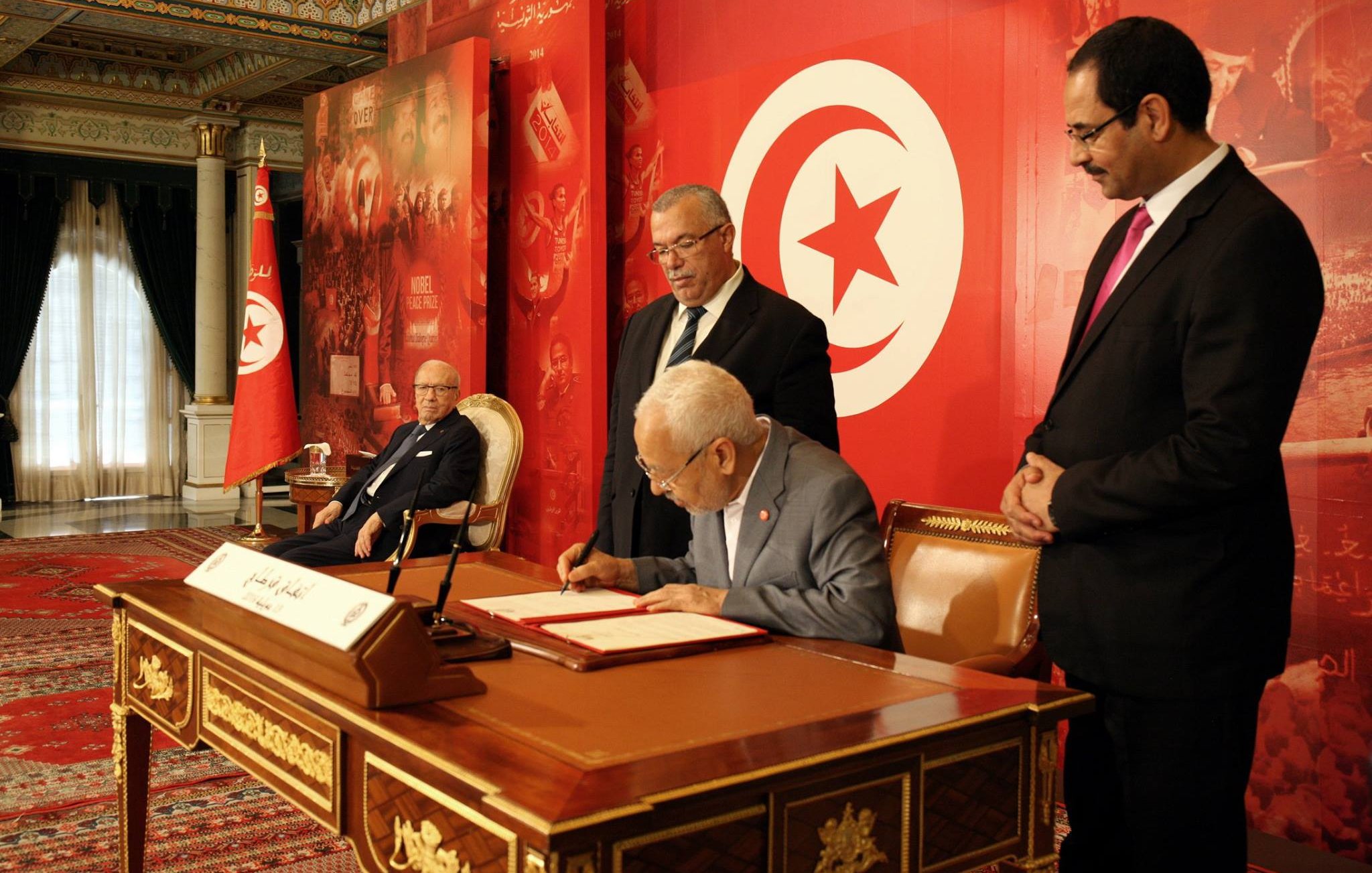 L'image montre une scène officielle où plusieurs personnes sont présentes dans un cadre prestigieux. Au premier plan, un homme assis à une table signe un document, tandis que d'autres hommes, debout à proximité, le regardent attentivement. En arrière-plan, on peut voir des drapeaux tunisiens et des éléments décoratifs évoquant une atmosphère solennelle. Le tout est agrémenté de couleurs vives, avec un fond rouge et des motifs qui semblent représenter des symboles nationaux.