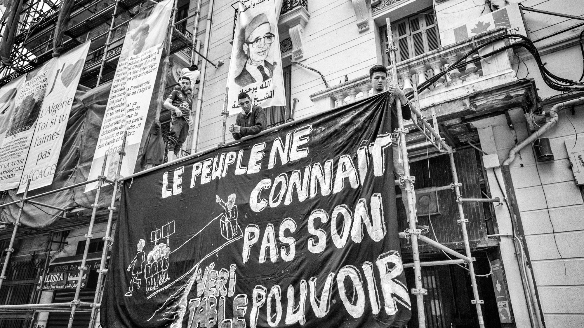 L'image présente une scène en noir et blanc où des personnes sont perchées sur un échafaudage face à un bâtiment. Ils tiennent une grande bannière sur laquelle est écrit en lettres blanches : "LE PEUPLE NE CONNAÎT PAS SON POUVOIR". En arrière-plan, on peut voir des affiches et des motifs typiques d'un environnement urbain. Cette composition évoque un sentiment de protestation ou de revendication sociale.