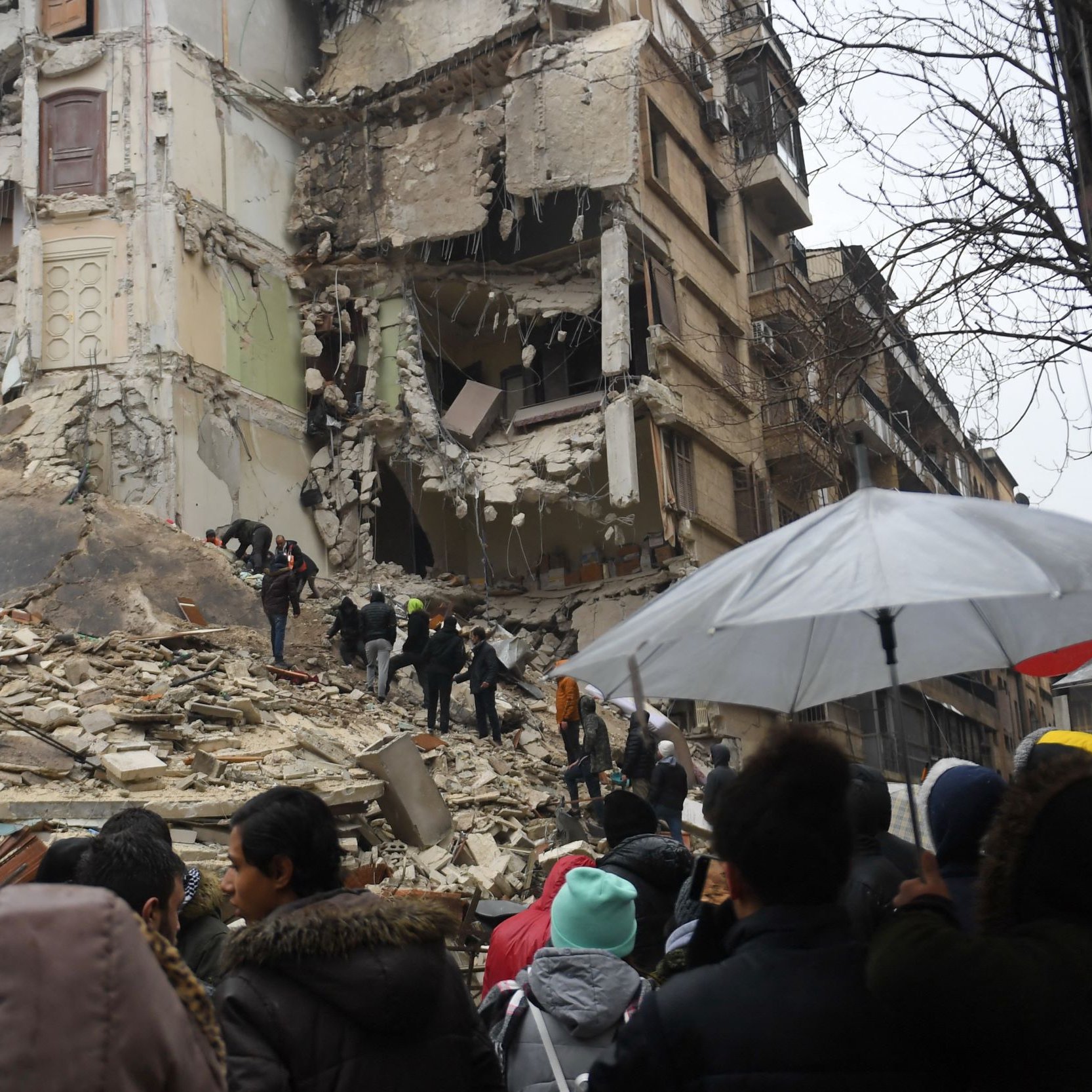 L'immagine mostra una scena dopo un terremoto devastante. Si può vedere un edificio gravemente danneggiato, con pareti crollate e macerie sparse. Alcuni soccorritori stanno cercando di recuperare persone intrappolate tra i detriti. Una folla di persone osserva da una certa distanza, alcune delle quali sono riparate sotto ombrelli, probabilmente a causa della pioggia. L'atmosfera è caratterizzata da tensione e preoccupazione, mentre le squadre di soccorso lavorano per salvare vite.