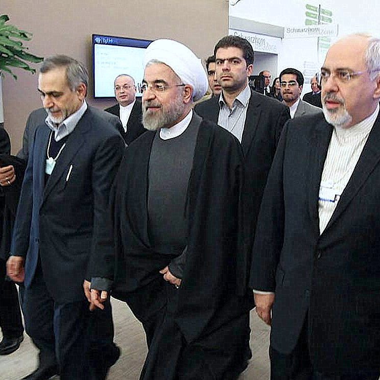 L'image montre un groupe d'hommes en costume formel, marchant ensemble dans un environnement apparemment professionnel. Certains d'entre eux portent des vêtements traditionnels, comme des chapeaux ou des manteaux, tandis que d'autres ont des costumes occidentaux. L'atmosphère semble être celle d'un événement officiel ou d'une conférence, avec des personnes à l'arrière-plan qui observent ou participent également. Leur expression indique une certaine concentration et un engagement dans la situation.