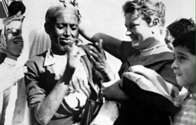 تظهر الصورة مجموعة من الأشخاص في احتفال يبدو أنه مليء بالحيوية والفرح. هناك شخص يظهر في المقدمة يرتدي ملابس تقليدية ويبدو أنه يشارك في عرض أو احتفال ثقافي. إلى جانبه، توجد سيدة تحمل ابتسامة على وجهها، مما يشير إلى جو من الألفة والود. الخلفية تحتوي على مزيد من الأشخاص الذين يظهرون في زوايا مختلفة، مما يعكس تفاعلًا اجتماعيًا نشيطًا. الألوان والتفاصيل تُسهم في الشعور بالبهجة والتفاهم بين الثقافات.