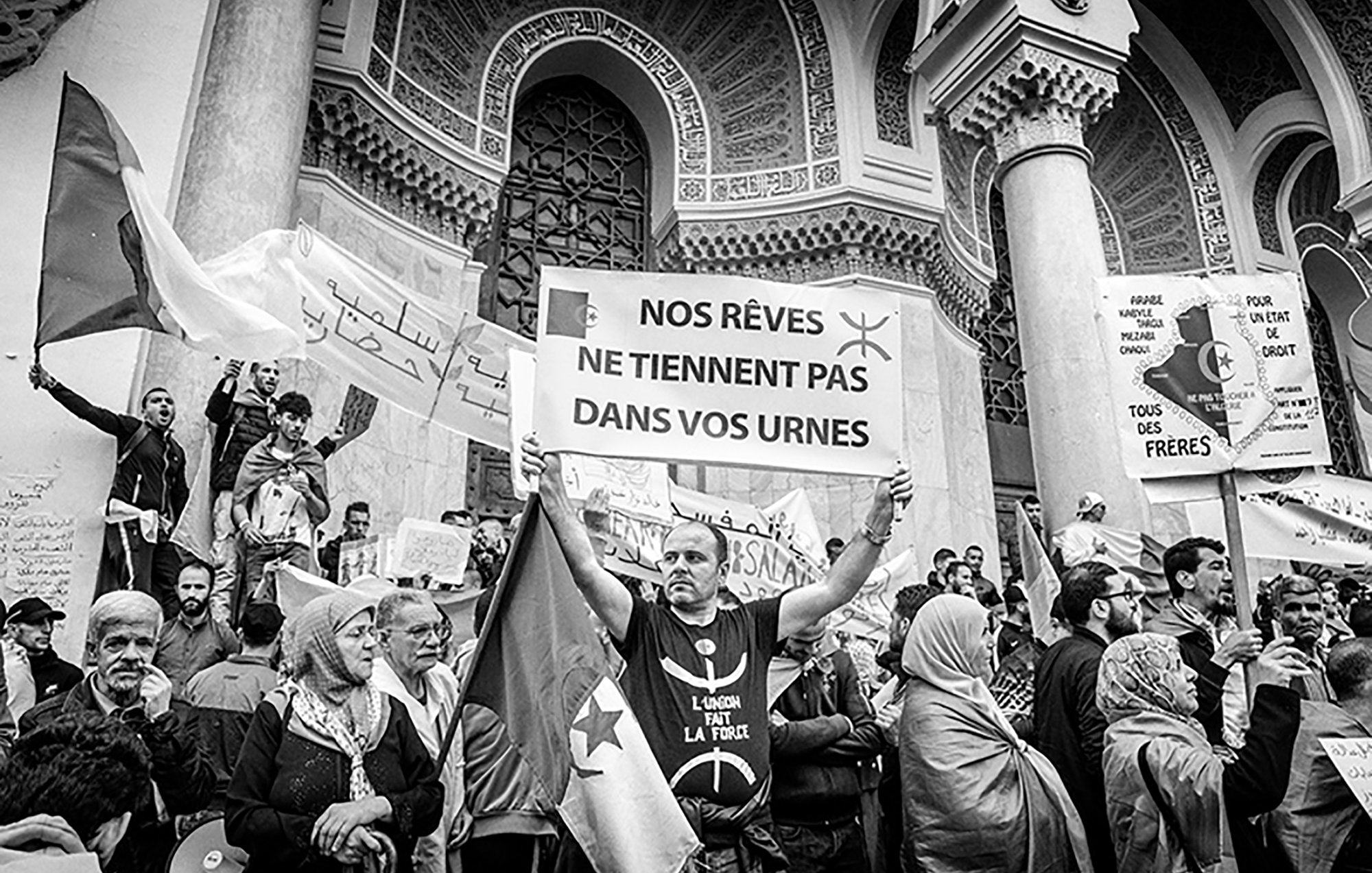 La imagen muestra una protesta o manifestación donde un grupo de personas sostiene pancartas y carteles. En el centro, un hombre sostiene un gran cartel con el mensaje "NOS RÊVES NE TIENNENT PAS DANS VOS URNES", que se traduce como "Nuestros sueños no caben en sus urnas". La escena está llena de gente, algunos con banderas y otros con rostros determinados, todos expresando descontento o reivindicaciones. El ambiente es de energía colectiva y compromiso social, y la imagen es en blanco y negro, lo que le da un tono más dramático y solemne.
