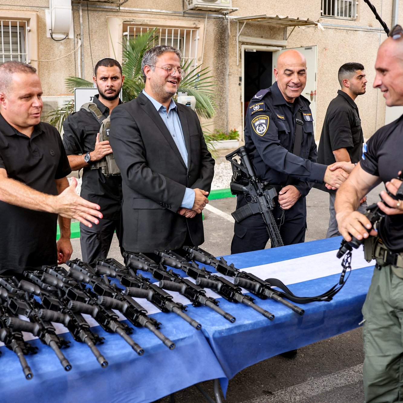 Un gruppo di uomini, alcuni in divisa, osserva fucili su un tavolo, mentre si stringono mani.