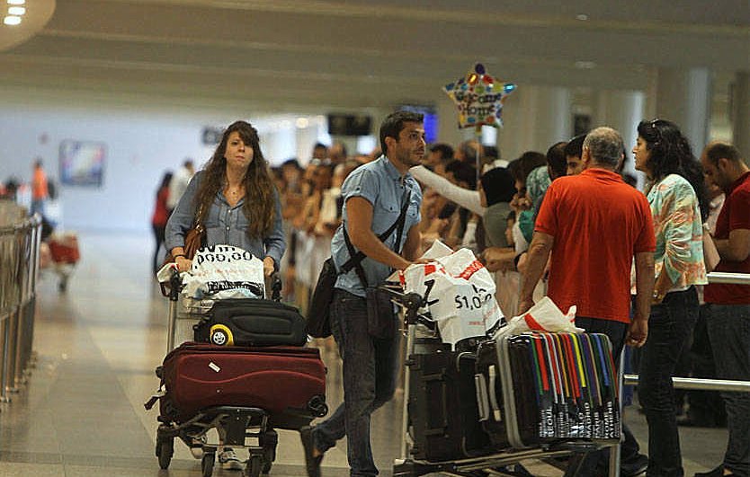 L'image montre une scène dans un aéroport, avec des voyageurs. À l'avant, un homme pousse une chariot de bagages et une femme derrière lui, également avec un chariot. Ils semblent attendent ou se déplacent parmi d'autres passagers. En arrière-plan, on aperçoit une longue file de personnes, probablement en attente de faire enregistrer leurs bagages ou de passer la sécurité. L'ambiance semble animée, typique des aéroports.