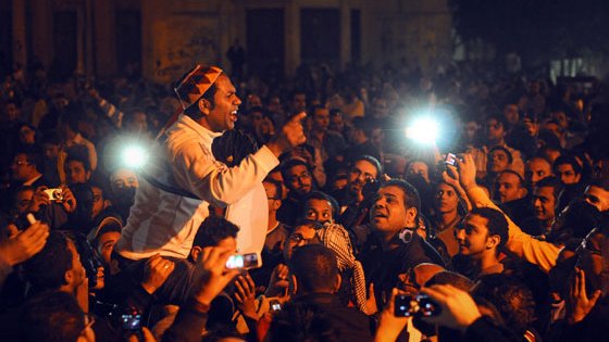 L'image montre une grande foule rassemblée dans une ambiance nocturne. Un homme au centre, visiblement passionné, est en train de parler ou de s'exprimer avec intensité. Les visages de la foule sont éclairés par des lumières, peut-être des téléphones portables ou des caméras. Les expressions des gens varient entre l'engagement, la curiosité et l'émotion, suggérant un moment de rassemblement ou de protestation. L'ensemble dégage une atmosphère vibrante et dynamique.