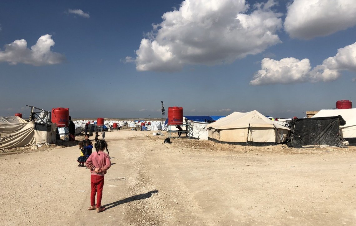 تظهر في الصورة مخيم للاجئين أو النازحين، حيث تتواجد عدة خيام مرتبة على جانبي الطريق. السماء صافية مع وجود سحب بيضاء، والأرض مرصوفة بالحجارة. يمكن رؤية بعض الأطفال يلعبون في المنطقة. يبدو أن المخيم يعاني من ظروف بسيطة، مما يشير إلى حياة موقتة للأشخاص المقيمين فيه.