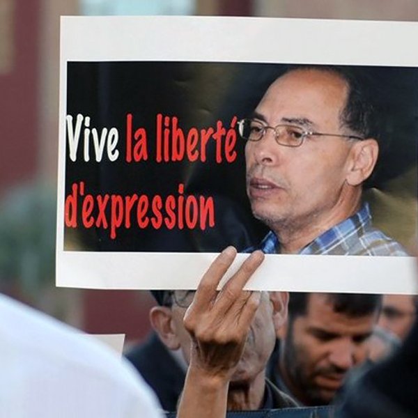 L'image montre une manifestation avec des personnes tenant des pancartes. L'une des pancartes porte le message « Vive la liberté d'expression » en rouge et noir, accompagnée d'une photo d'un homme. L'atmosphère semble engagée et déterminée, reflétant une revendication pour la liberté d'expression. Les manifestants semblent concentrés sur cette cause.