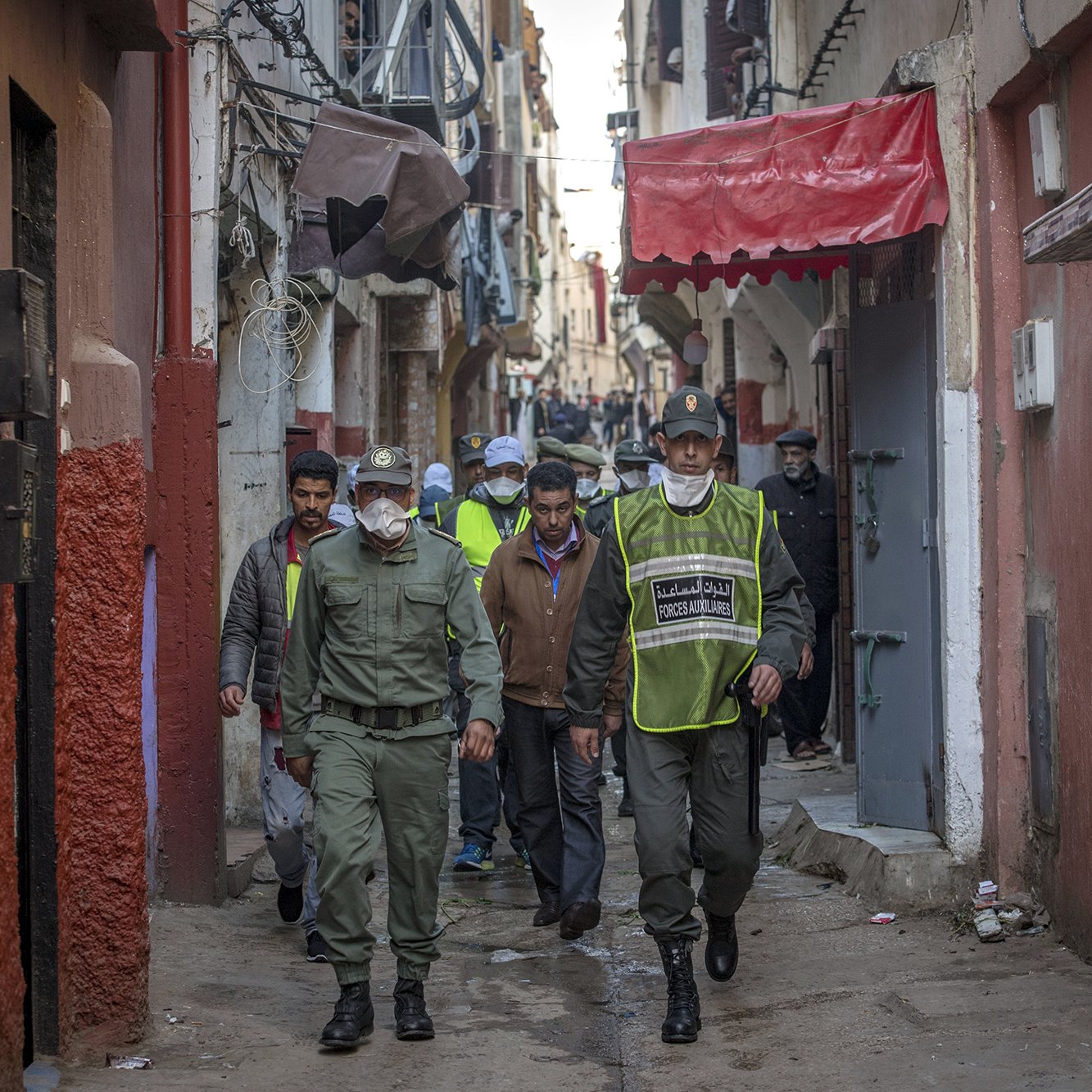 L'image montre une ruelle étroite et animée, typique d'une ville marocaine. Au premier plan, des agents de police ou des membres de la sécurité patrouillent. Ils portent des uniformes et des masques, indiquant une attention à la sécurité ou à la santé publique. En arrière-plan, on peut voir des habitants et d'autres personnes dans la rue, ainsi que des bâtiments aux façades colorées. L'atmosphère semble à la fois dynamique et surveillée, reflétant la vie quotidienne dans ce type d'environnement urbain.