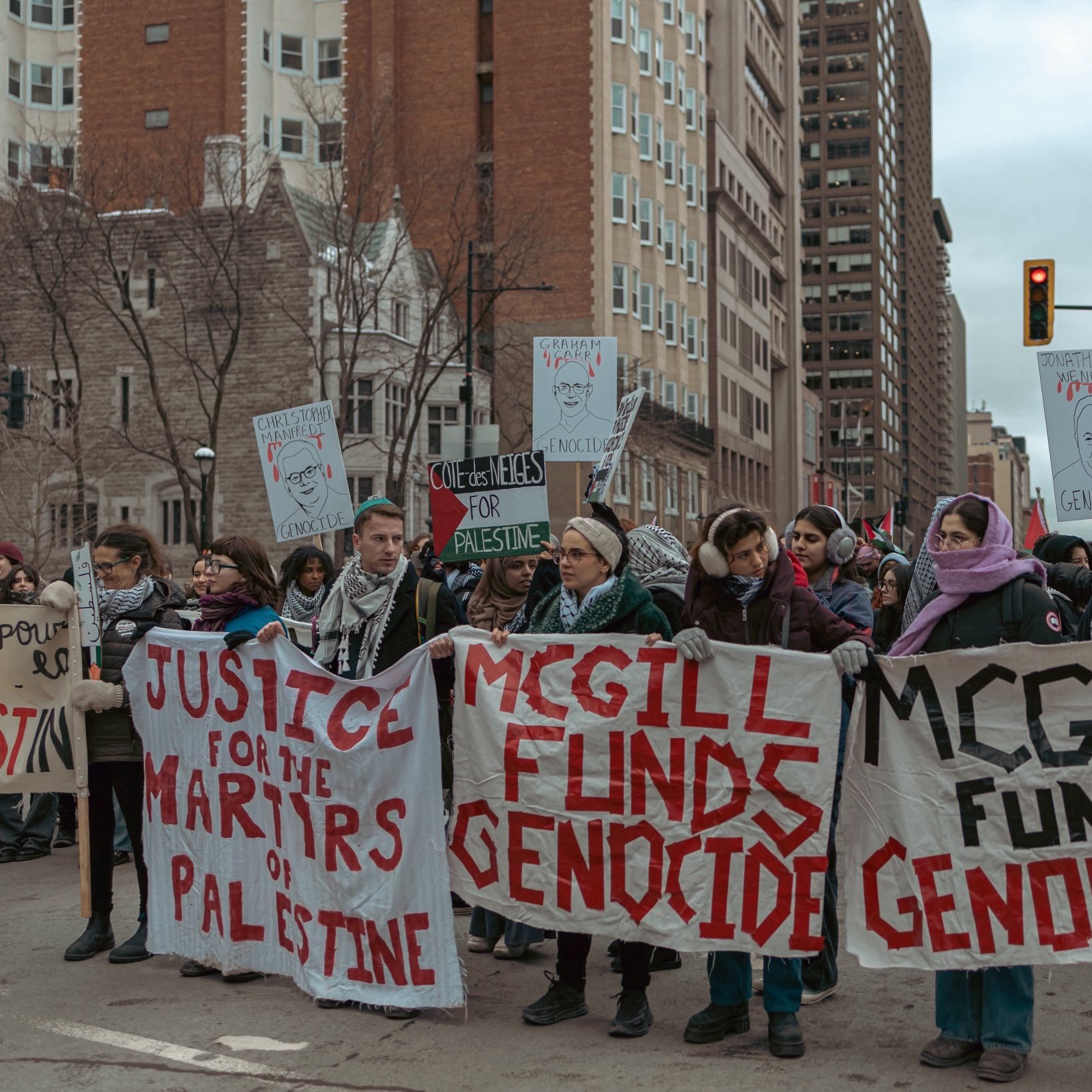 The image depicts a group of protesters marching in a city setting. They are holding banners and signs with messages emphasizing justice for Palestine and accusing McGill University of funding genocide. The atmosphere appears serious and focused, with the protesters displaying solidarity on the issue. The urban environment features tall buildings in the background, indicating that this is a public demonstration.