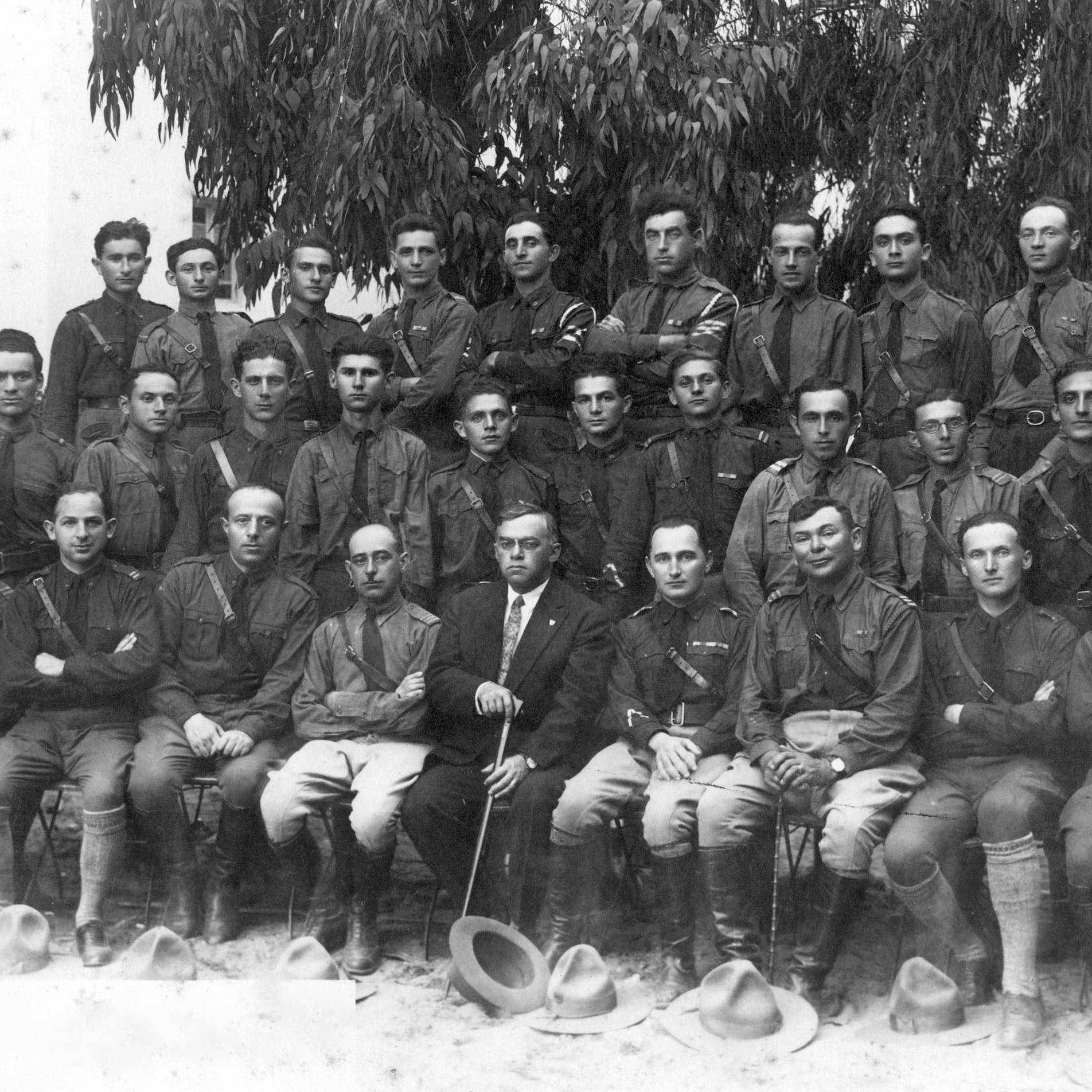 L'image montre un groupe de soldats en uniforme, probablement pris au début du 20e siècle. Ils sont assis et debout en rangée, avec des expressions sérieuses. Au centre, un homme en costume, tenant une canne, semble superviser le groupe. L'arrière-plan présente des arbres feuillus, ajoutant une touche naturelle à la scène. L'ambiance générale évoque une certaine solennité et un sentiment de camaraderie parmi les membres du groupe.