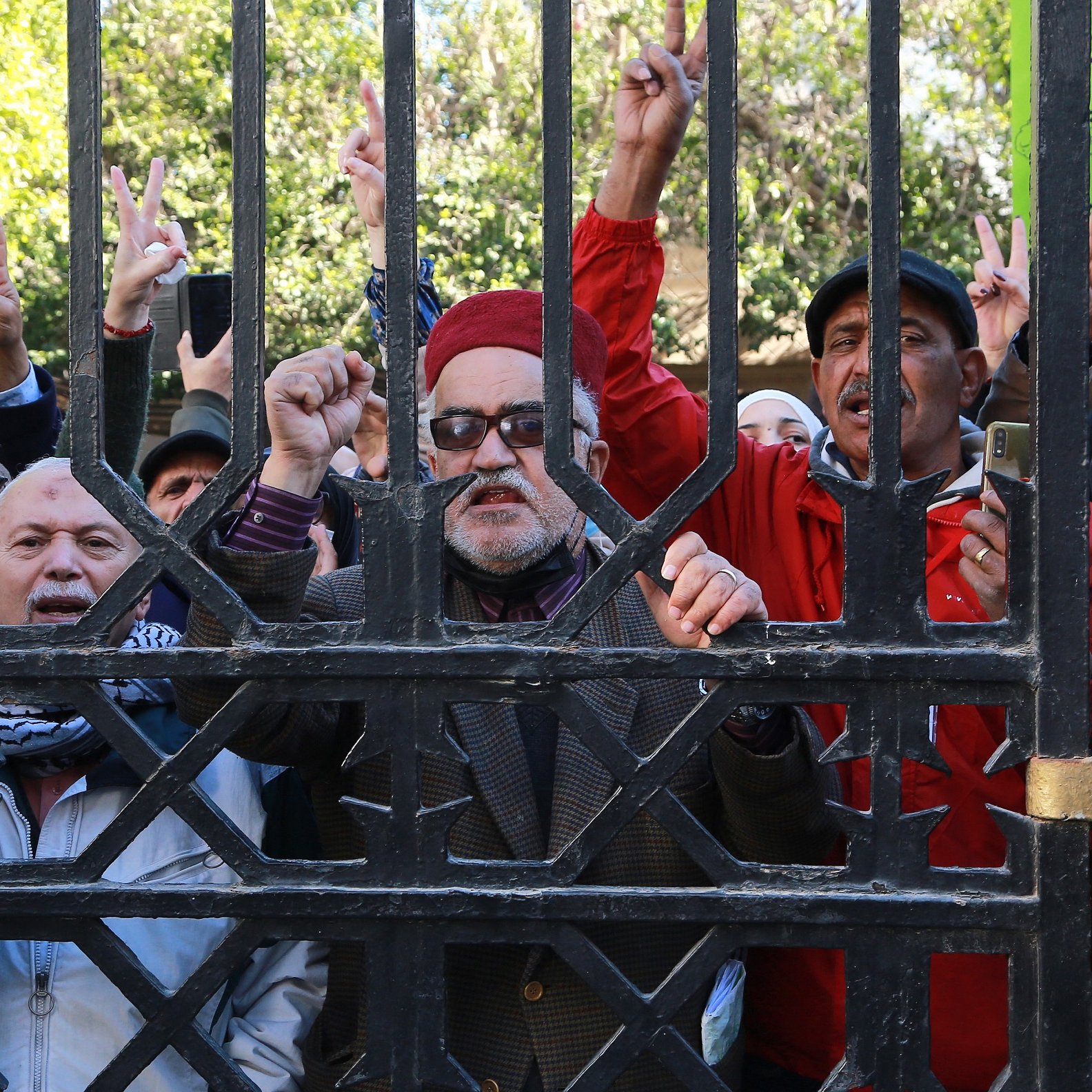 L'image montre un groupe de personnes rassemblées derrière une grille. Certains individus lèvent leurs doigts en forme de V, symbole de la paix ou de la victoire. Les expressions des participants varient, allant de la détermination à l'enthousiasme. Il semble qu'ils soient engagés dans une manifestation ou un rassemblement. Les arrière-plans sont flous, mettant l'accent sur les manifestants et leur activité.