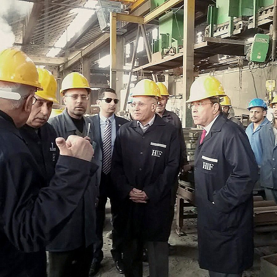 L'image montre un groupe de personnes dans un environnement industriel, probablement dans une usine. Les participants portent des casques de sécurité jaunes, ce qui indique qu'ils sont dans un lieu de travail où des précautions de sécurité sont nécessaires. Certains hommes sont en train de discuter ou d'écouter attentivement, tandis que d'autres observent la scène. En arrière-plan, on peut voir des machines et des équipements typiques d'un environnement de production. L'atmosphère semble être sérieuse et professionnelle.