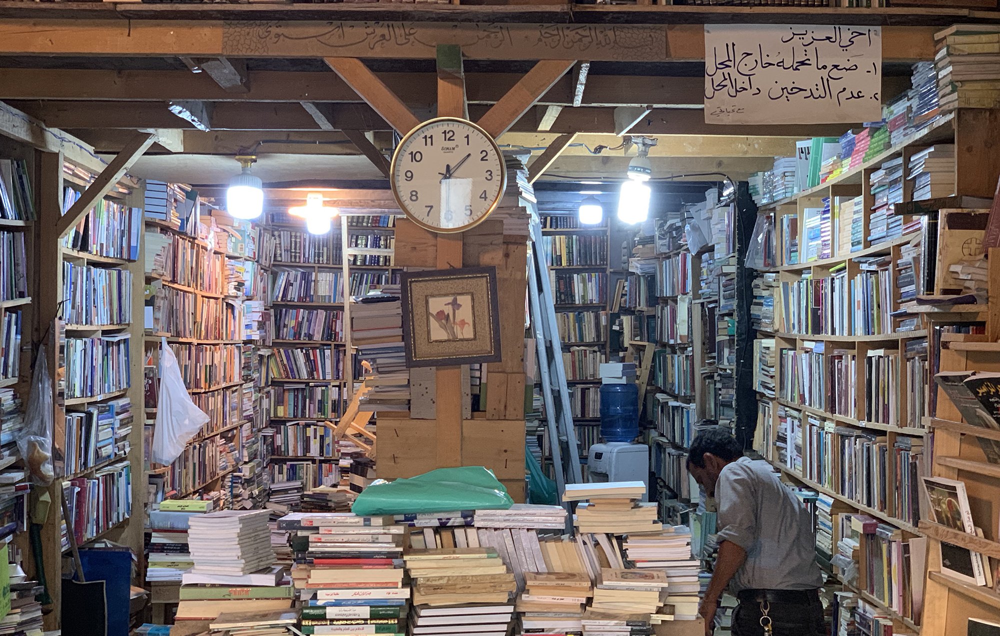 تظهر الصورة مشهداً داخلياً لمكتبة قديمة. تتراص الكتب على الرفوف الخشبية بشكل كثيف، مع ظهور بعض الكتب المكدسة على الأرض. في منتصف الصورة، يوجد ساعة حائط تشير إلى الوقت. كما يوجد شخص في الجهة اليمنى من الصورة، يبدو أنه يتفحص الكتب. الأضواء تتدلى من السقف، مما يضفي جواً دافئاً على المكان. في الأعلى، توجد لافتة مكتوبة توضح معاني تتعلق بالمكتبة.