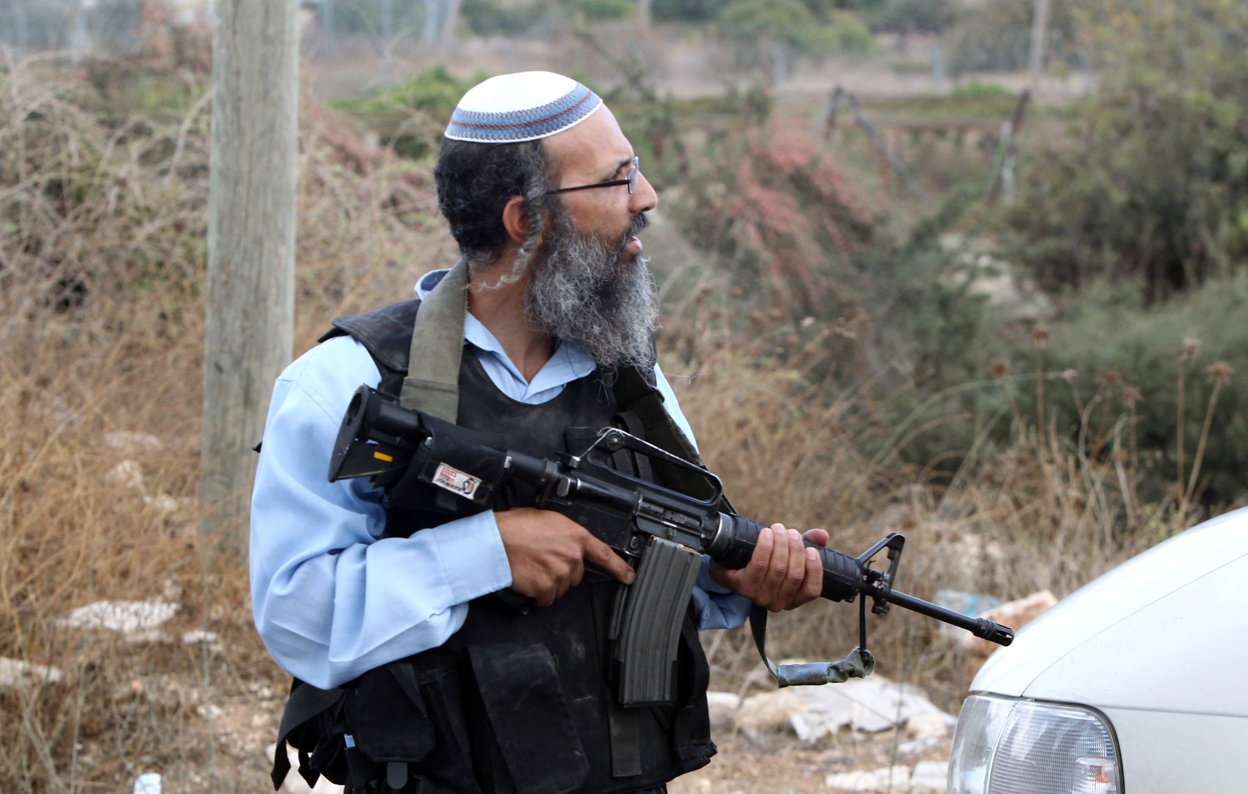 L'image montre un homme qui porte une kippa et une veste de protection. Il est armé d'un fusil automatique et se tient près d'une voiture dans un environnement extérieur, avec de la végétation et des débris autour de lui. Son expression et sa posture suggèrent qu'il est en alerte ou surveille les alentours.