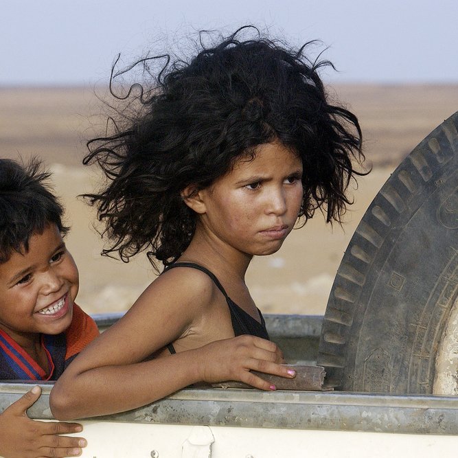L'image montre deux enfants, un garçon et une fille, sur un véhicule. Le garçon, souriant, a les cheveux noirs et bouclés, et il semble joyeux. La fille, dont les cheveux sont également très volumineux, a une expression sérieuse ou pensive. Ils sont entourés d'un paysage désertique aux nuances de jaune et de beige. L'arrière-plan suggère un environnement rustique et chaud.