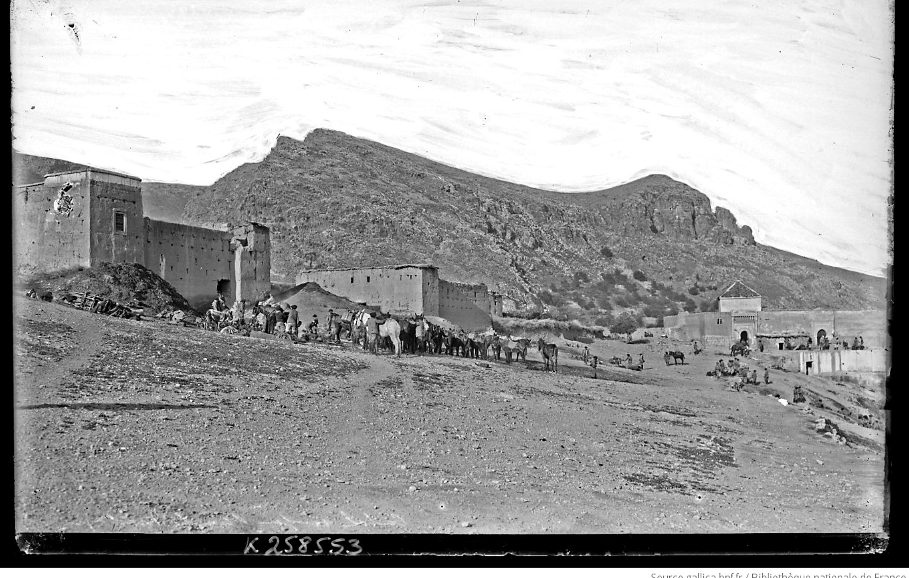 L'image présente un paysage désertique avec des montagnes en arrière-plan. À gauche, on aperçoit des bâtiments en pierre, probablement des fortifications ou des maisons, qui semblent très anciens. Au premier plan, un groupe de personnes est visible, accompagné de plusieurs animaux de charge, comme des chameaux ou des ânes. Le ciel est clair, montrant une atmosphère paisible, et le terrain est aride, typique des régions montagneuses désertiques. L'ensemble dégage une impression d'histoire et de culture.