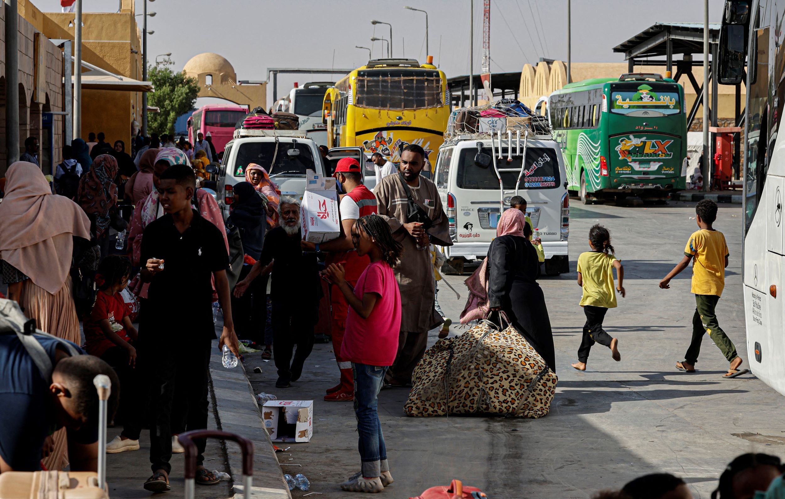 در تصویر، یک ایستگاه اتوبوس یا ترمینال مشاهده می‌شود که در آن تعدادی اتوبوس در حال توقف هستند. مردم زیادی در حال رفت و آمد هستند، برخی در حال حمل چمدان و بارهای خود هستند. در میان این افراد، کودکان نیز دیده می‌شوند که در حال بازی یا حرکت هستند. برخی از افراد در حال استراحت و نشستن بر روی ساک‌های خود هستند. فضای ترمینال به نظر شلوغ و پرجنب و جوش می‌آید و آفتاب روشن در حال تابش است.