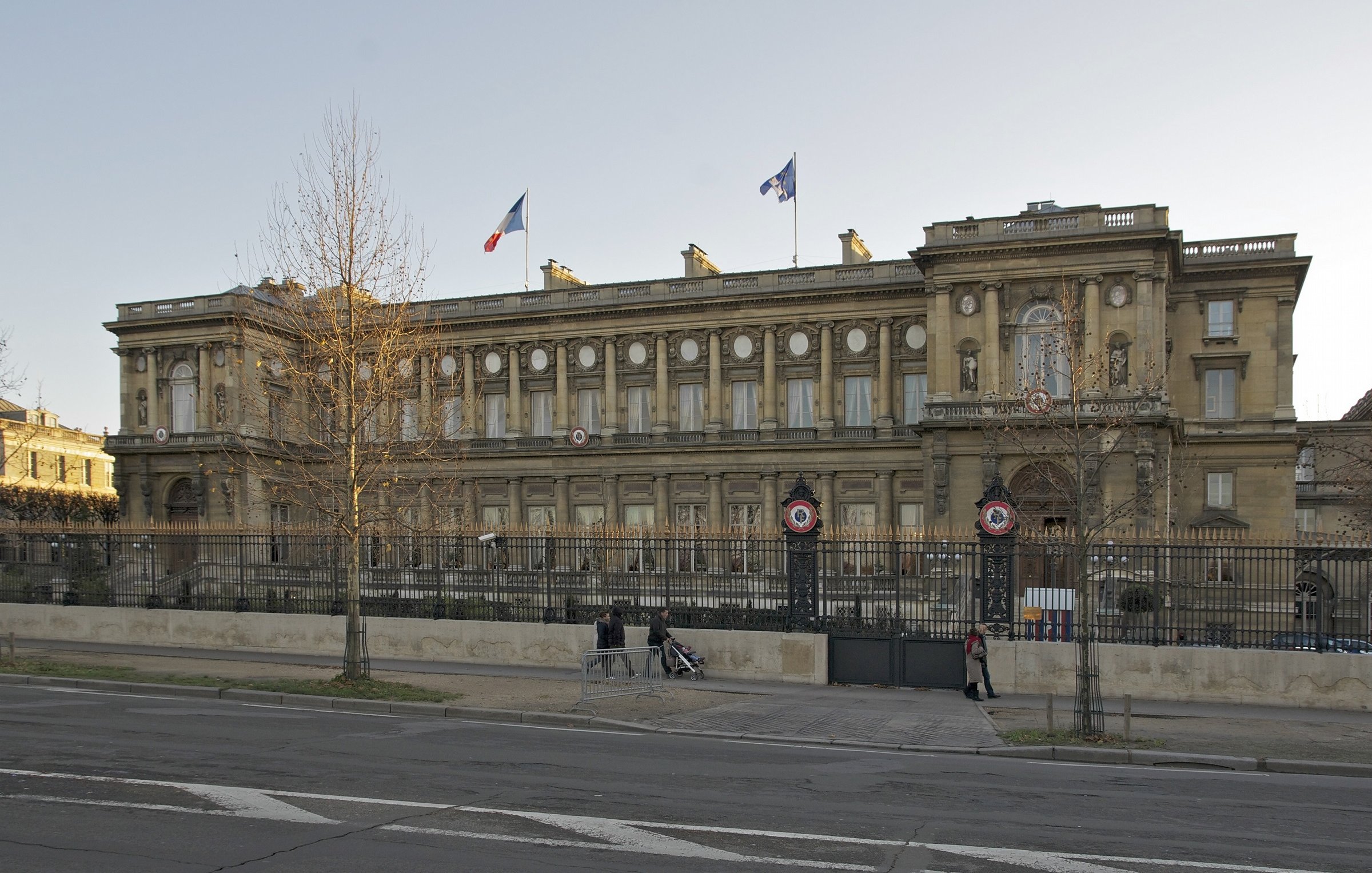 L'image montre un bâtiment imposant avec une architecture classique, situé dans un espace urbain. On peut voir des drapeaux français flottant au sommet, et le bâtiment est entouré d'une grille. La façade est riche en détails, avec des colonnes et des ornements décoratifs. Il semble être un édifice gouvernemental ou historique, situé probablement dans une grande ville française. Des arbres sont visibles sur les côtés, et une route passe devant le bâtiment.