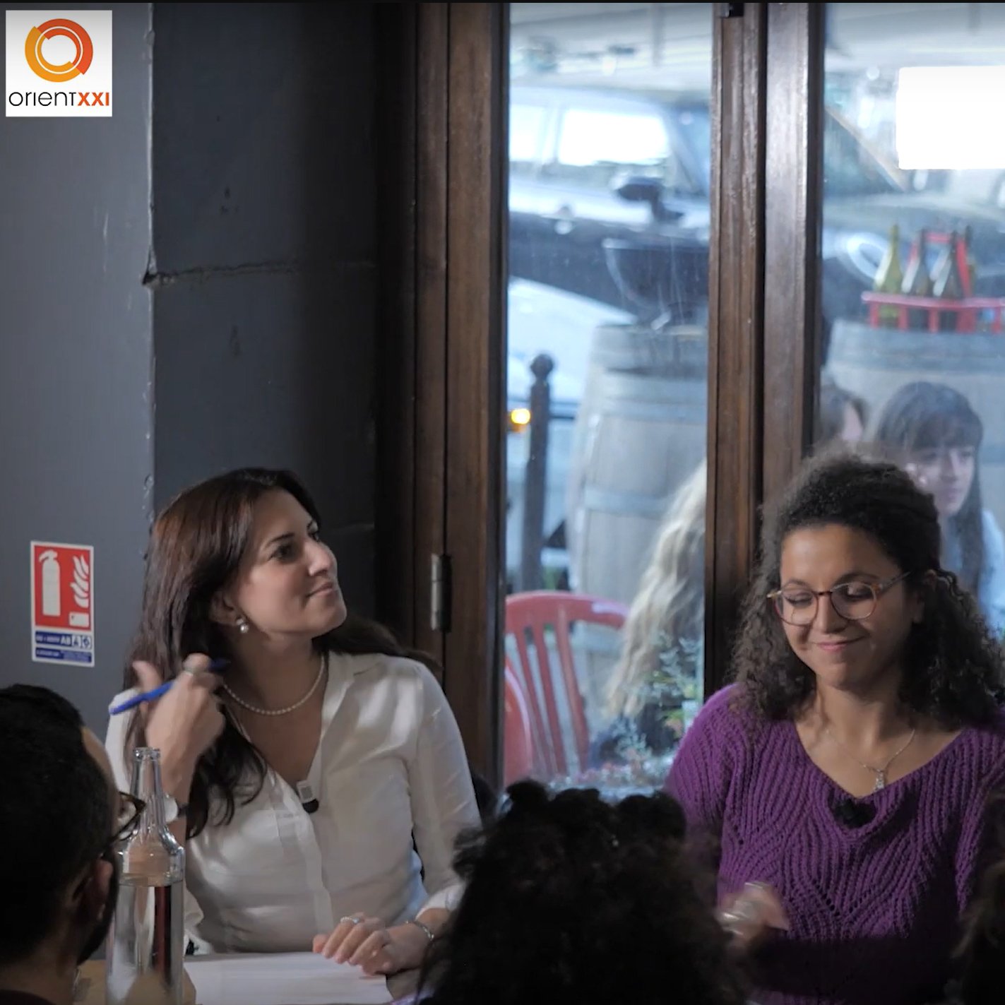 L'image montre une scène intérieure où plusieurs personnes sont assises à des tables. Une femme se tient debout au premier plan, tenant un microphone, probablement en train de parler ou de présenter quelque chose. Elle porte des vêtements noirs et semble engagée dans une conversation dynamique. À sa gauche, une autre femme sourit, tandis qu'à sa droite, une femme avec des lunettes apporte une expression de concentration. À l'arrière-plan, on peut voir des clients assis, créant une atmosphère animée. Les grandes fenêtres laissent entrer la lumière et montrent une vue partielle de l'extérieur.