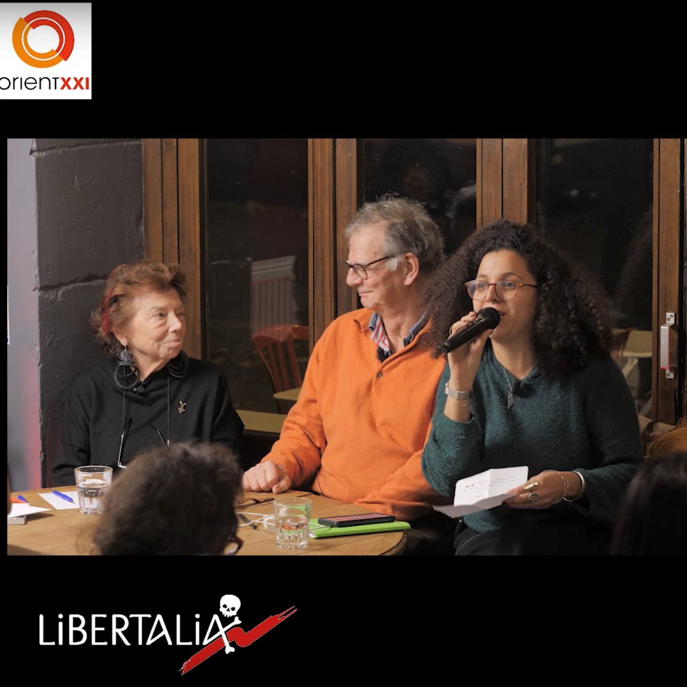 L'image montre un événement en cours avec des participants assis autour d'une table. À gauche, un homme et une femme discutent, la femme tenant un micro et s'adressant à l'audience. L'homme à ses côtés sourit et semble attentif. À droite, il y a une couverture d'un livre intitulé "Journal de bord de Gaza", avec des éléments graphiques stylisés. Le tout évoque une ambiance conviviale et engagée, orientée vers la discussion d'une œuvre littéraire ou d'une thématique importante.