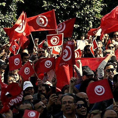 L'image montre une foule de personnes rassemblées, agitant des drapeaux tunisiens rouges avec un cercle blanc et une étoile à cinq branches. Les manifestants semblent animés, levant les bras en signe de solidarité ou de protestation. On peut voir un paysage urbain en arrière-plan avec des arbres, ce qui suggère un rassemblement en plein air. L'atmosphère semble énergique et engagée, reflétant un moment de mobilisation collective.