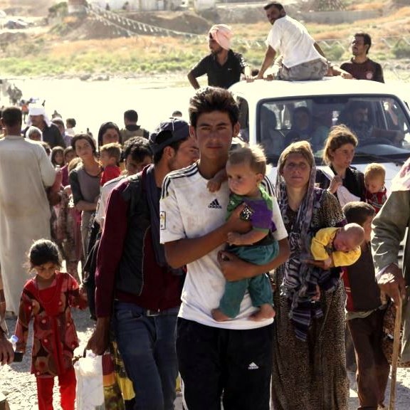 تظهر الصورة مجموعة من الأشخاص الذين يبدون وكأنهم في حالة نزوح. يتواجدون في منطقة مفتوحة، حيث يمشي البعض على الطريق بينما يقف آخرون بالقرب من سيارات. هناك أطفال يحملهم بعض البالغين، مما يعكس معاناة وتحديات العائلات في ظل ظروف صعبة. تُظهر الصورة أيضاً تنوعاً في الأعمار والملابس، مما يدل على أن هؤلاء الأشخاص قد يأتون من خلفيات مختلفة. الأجواء تبدو حزينة وتعبر عن الشعور بالفقد والبحث عن الأمان.