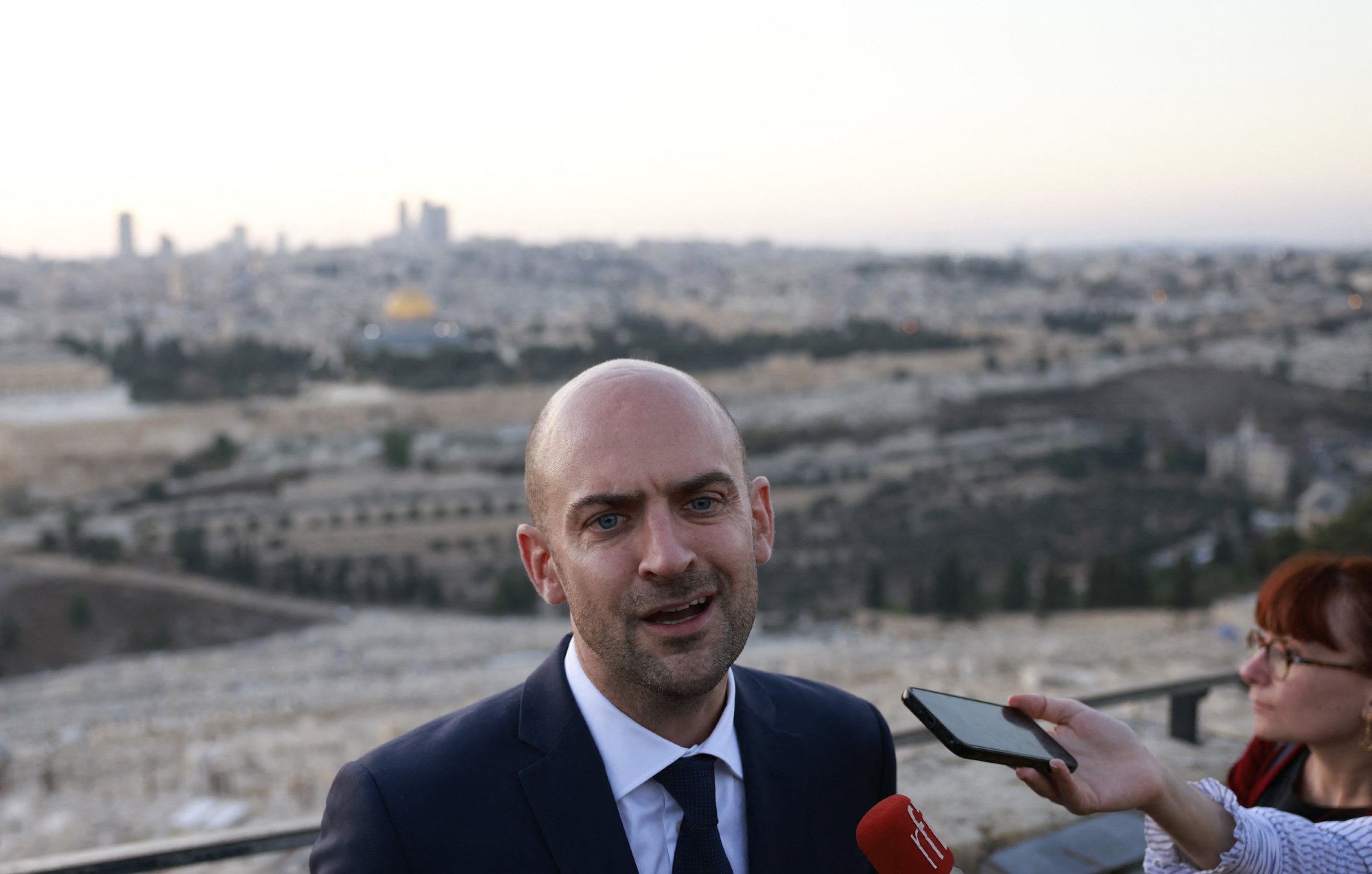 در این تصویر، یک مرد با سر طاس و کت و شلوار تیره در حال صحبت به دوربین و خبرنگاران است. او در یک مکان آزاد ایستاده و پشتش چشم‌انداز شهر قابل مشاهده است. درختان و ساختمان‌ها در دوردست دیده می‌شوند، و به نظر می‌رسد که این تصویر در یک زمان غروب یا صبح زود گرفته شده است. خبرنگاری نیز در سمت راست او قرار دارد و در حال ثبت‌کردن یا پرسش کردن از اوست.