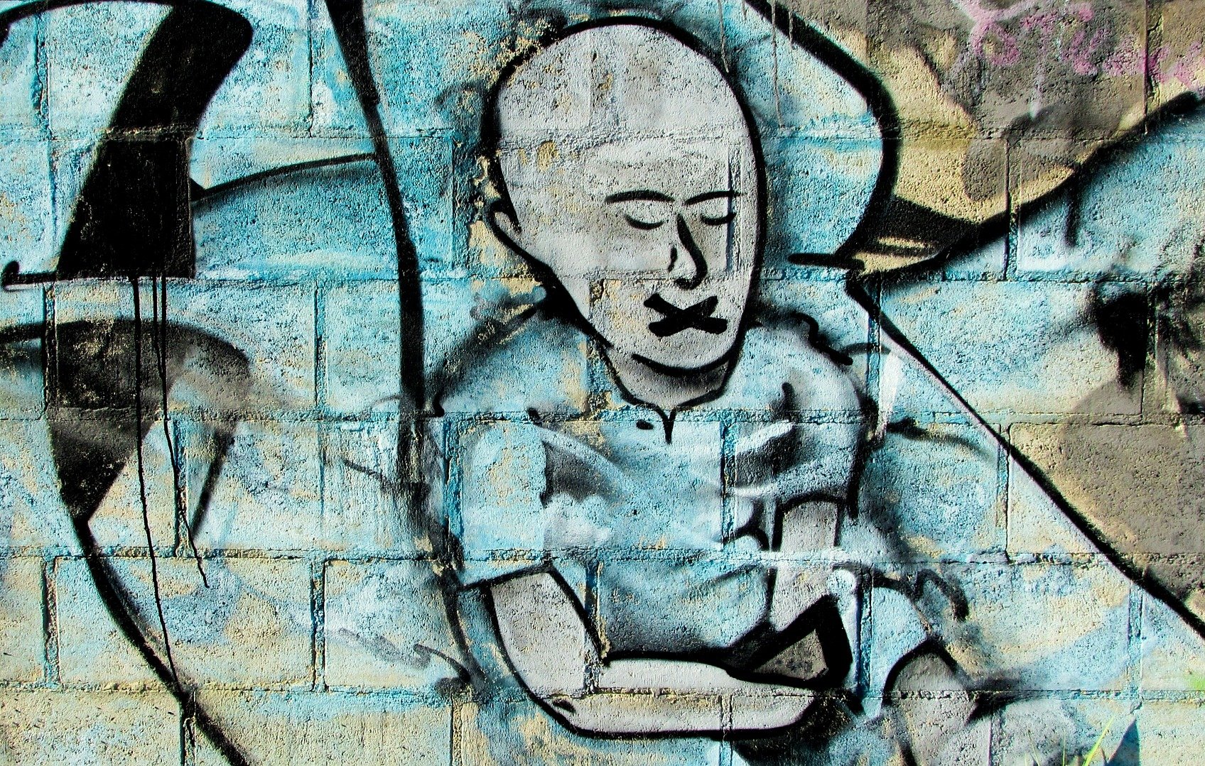 الصورة تظهر جدارية على جدار من الطوب، حيث تُصور رسماً لشخص يجلس مع ذراعيه على ركبتيه. الشخص يبدو بلا شعر وله تعبير هادئ، وفمه مُكمم، مما يُعطي انطباعاً بأن هناك رسالة تتعلق بالصمت أو القمع. الخلفية تحتوي على تدرجات من اللون الأزرق، مما يُضفي بعداً جمالياً على العمل الفني. الجدارية تعكس أسلوب فن الشارع الذي يُستخدم للتعبير عن القضايا الاجتماعية أو السياسية.