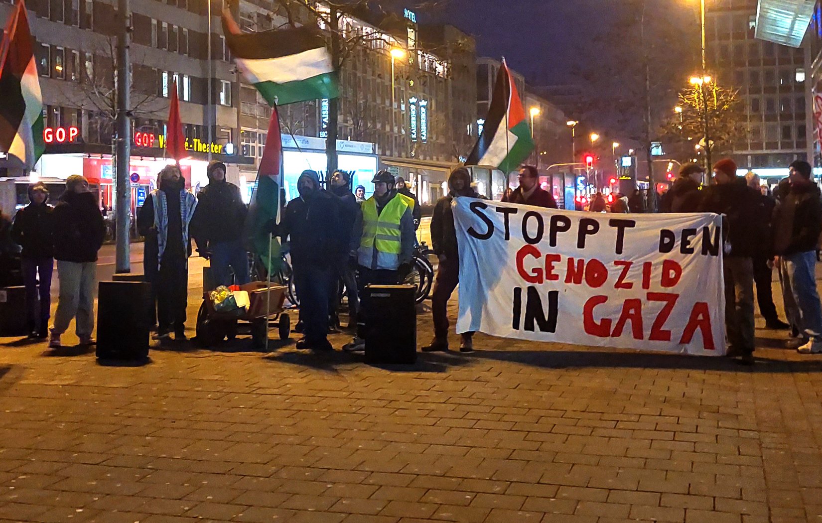 تُظهر الصورة مجموعة من المتظاهرين في مكان عام، يحملون لافتة كبيرة مكتوب عليها "STOPPT DEN GENOZID IN GAZA"، مما يعني "أوقفوا الإبادة الجماعية في غزة" باللغة الألمانية. يُلاحظ في الصورة أيضًا العلم الفلسطيني، حيث يرفع بعض المتظاهرين الأعلام دعمًا للقضية. يبدو أن التظاهرة تُعقد في وقت متأخر من اليوم، حيث تُضاء الأضواء في الشارع المحيط.