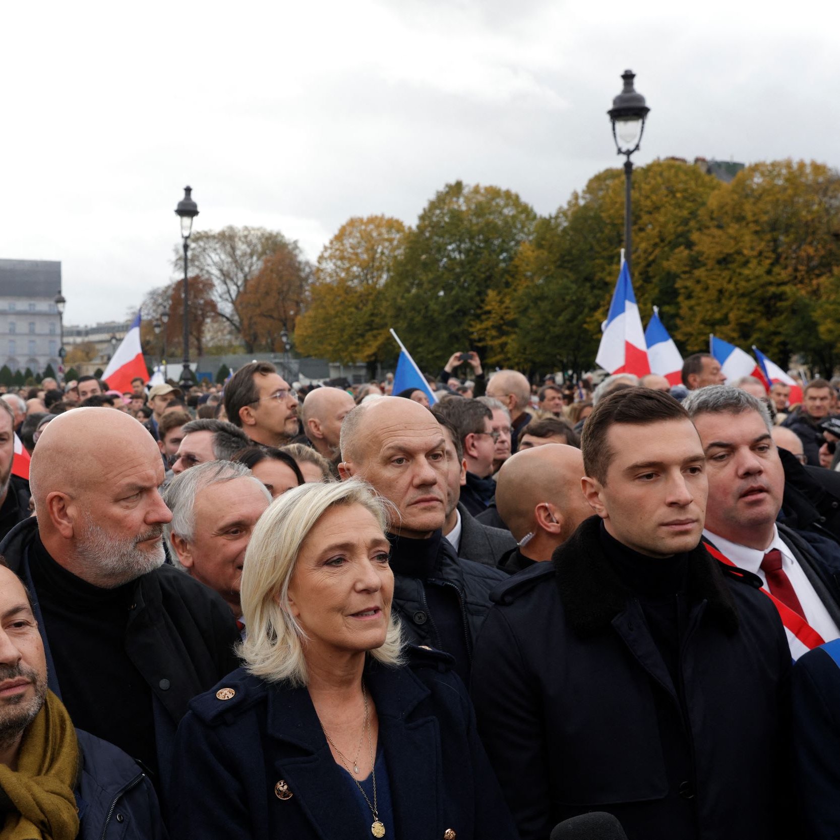 در این تصویر، گروهی از مردم در یک تجمع یا راهپیمایی در فضای باز به نمایش درآمده‌اند. بسیاری از افراد پرچم‌های فرانسه را در دست دارند. در میان جمعیت، افرادی با چهره‌های مختلف دیده می‌شوند که نشان‌دهنده نظرات و احساسات مختلفی هستند. برخی از افراد لباس‌های رسمی و گرم پوشیده‌اند و به نظر می‌رسد موضوع این تجمع سیاسی باشد. آسمان ابری است و فضای عمومی کمی شلوغ و پرتحرک به نظر می‌رسد.