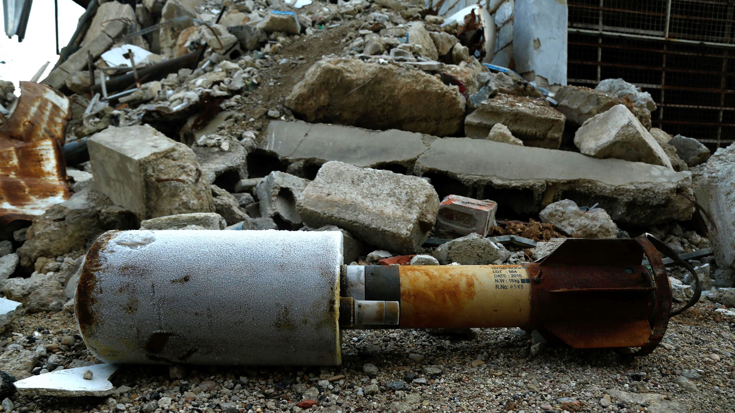 The image shows a missile or bomb lying on the ground among rubble and debris. The surrounding area is chaotic, with broken concrete and scattered materials suggesting destruction, possibly from an explosion or military action. The missile itself appears weathered and is partially covered in grime, indicating it has been in this location for some time.
