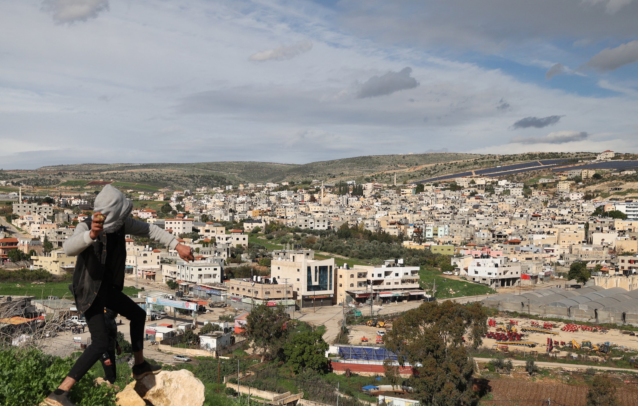 L'image montre une vue panoramique d'un paysage urbain avec de nombreux bâtiments construits, probablement dans une région montagneuse ou vallonnée. Au premier plan, une personne est en train de sauter ou de se déplacer sur des rochers. L'horizon est marqué par des collines et des nuages, et on peut voir des routes et des structures en dessous, indiquant une zone habitée. La scène dégage une atmosphère dynamique, avec un mélange d'éléments naturels et urbains.