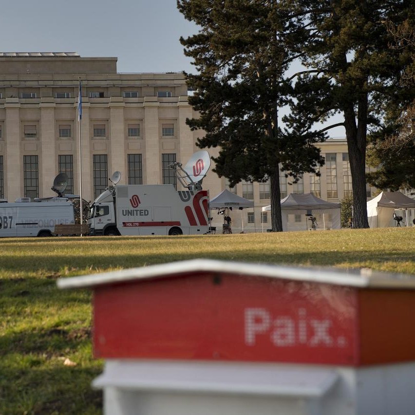 L'image montre un bâtiment imposant, probablement un palais ou un bâtiment gouvernemental, situé à Genève. À l'avant-plan, on aperçoit une petite structure rouge portant le mot "Paix". En arrière-plan, on distingue des véhicules de télévision et des tentes, indiquant qu'un événement ou une conférence pourrait avoir lieu. Le cadre est agrémenté d'arbres, suggérant un espace extérieur agréable.
